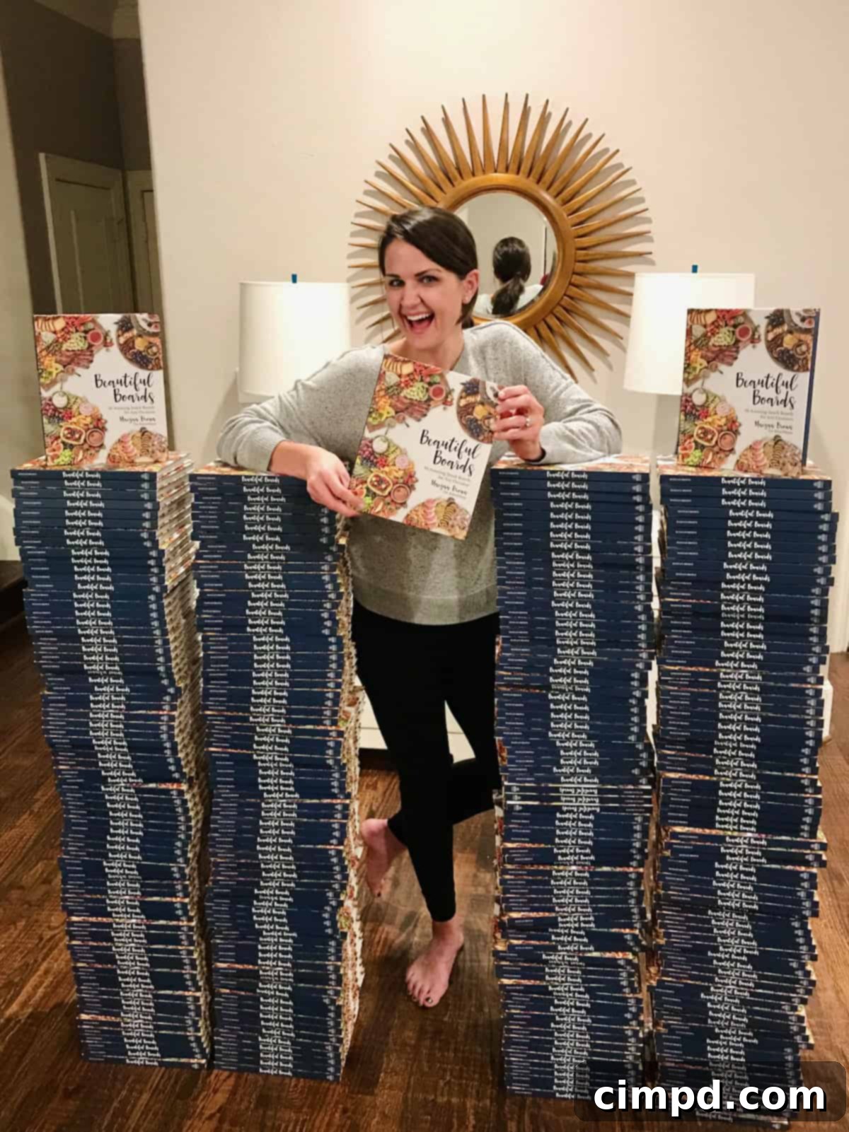 Maegan Brown stands proudly next to three towering stacks of her popular cookbook, Beautiful Boards, showcasing its success.