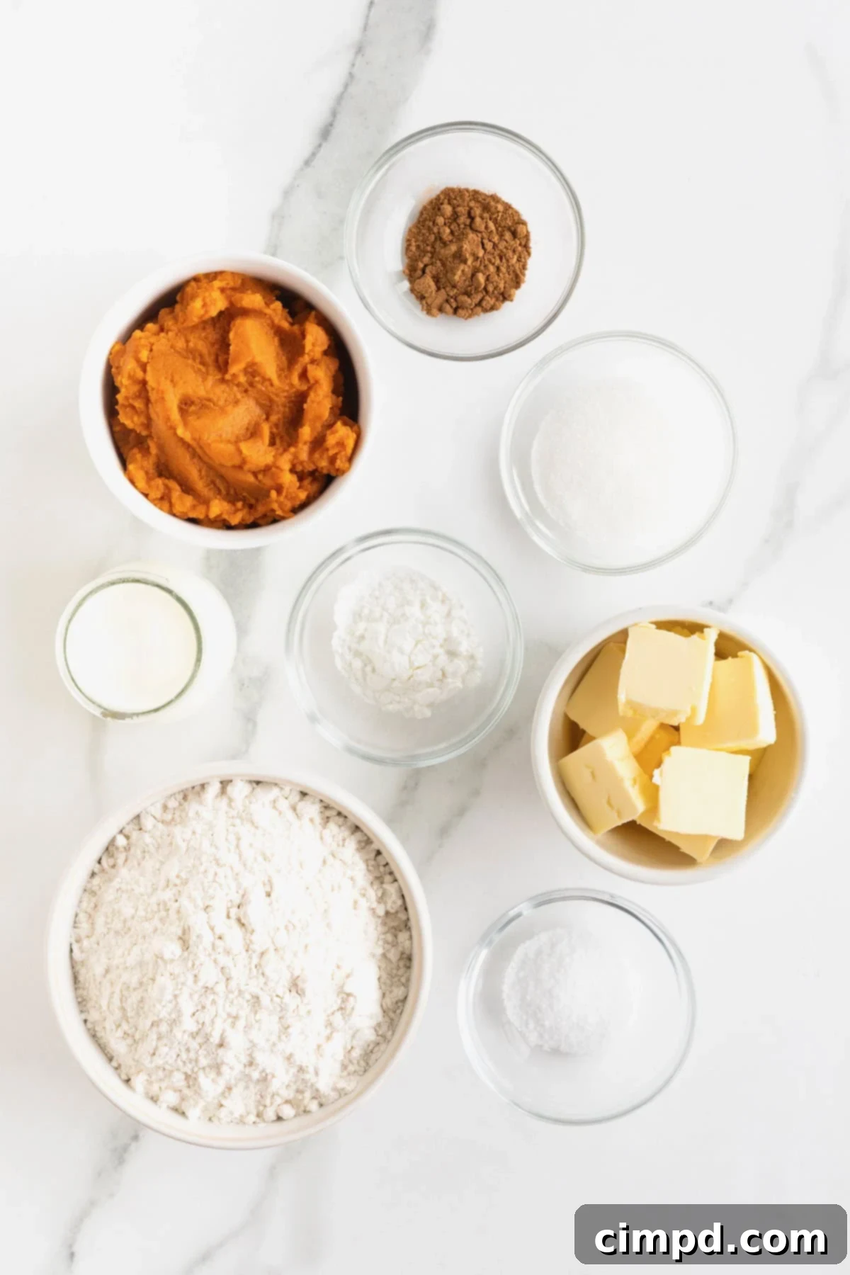 Ingredients to make pumpkin biscuits, including flour, sugar, spices, butter, milk, and pumpkin puree, arranged in small glass dishes on a white marble counter.