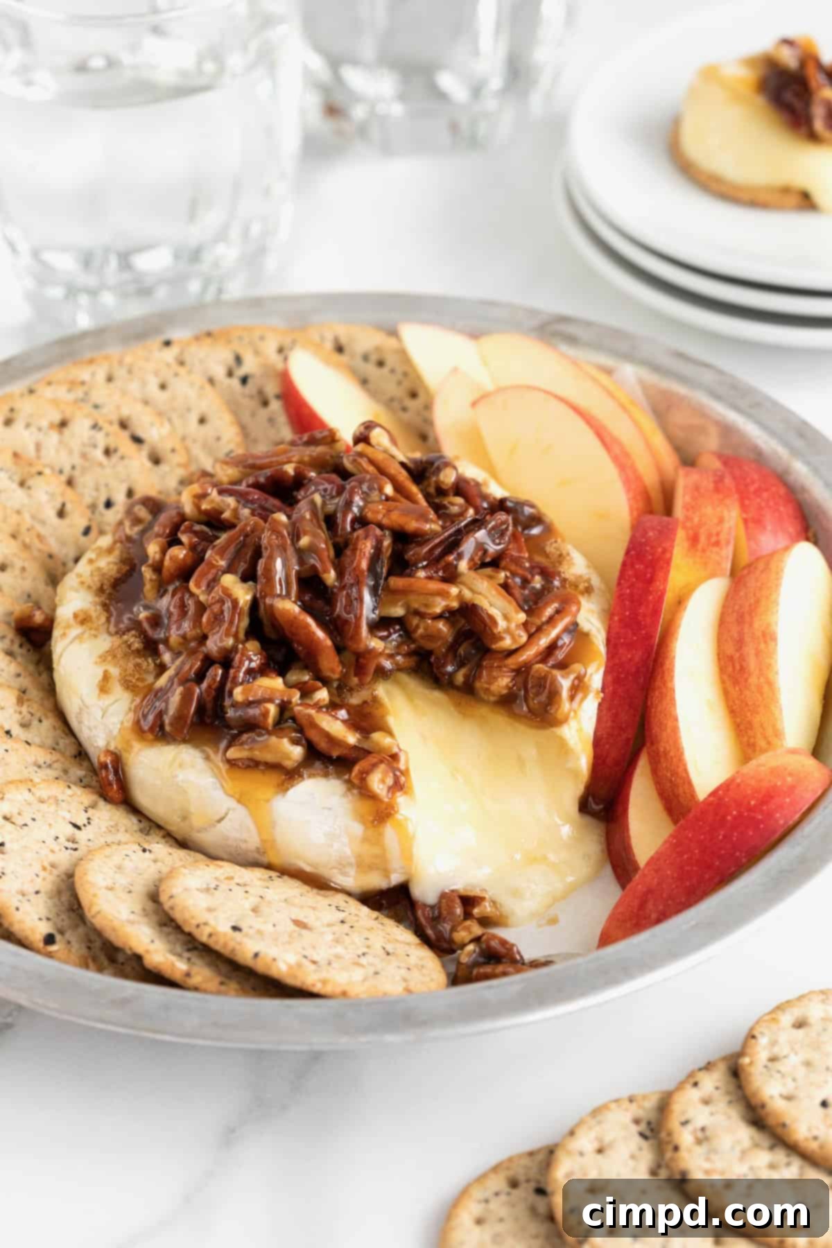 Caramel Pecan Baked Brie 2 A round aluminum baking dish with a wheel of brie in the middle, and apple slices and water crackers around the outside. The brie is topped with a pecan sauce.