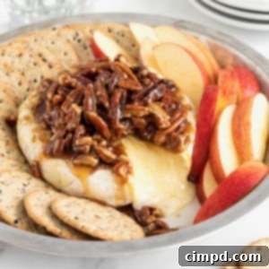 Caramel Pecan Baked Brie 10 A round aluminum baking dish with a wheel of brie in the middle, and apple slices and water crackers around the outside. The brie is topped with a pecan sauce.