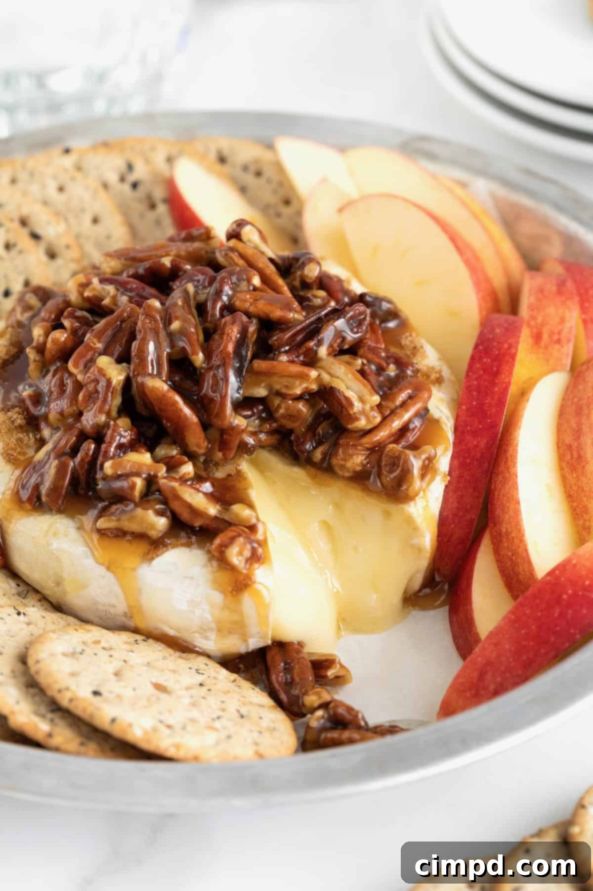 Caramel Pecan Baked Brie 4 A round aluminum baking dish with a wheel of brie in the middle, and apple slices and water crackers around the outside. The brie is topped with a pecan sauce.