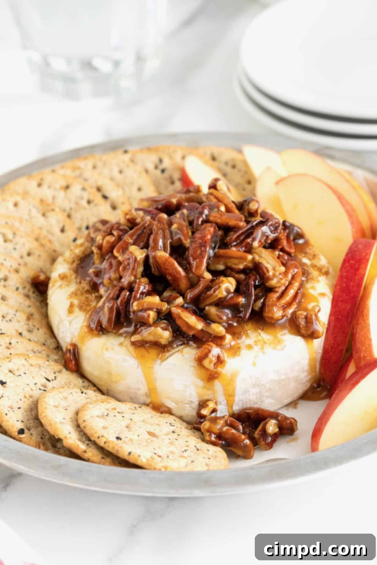Caramel Pecan Baked Brie 9 A round aluminum baking dish with a wheel of brie in the middle, and apple slices and water crackers around the outside. The brie is topped with a pecan sauce.