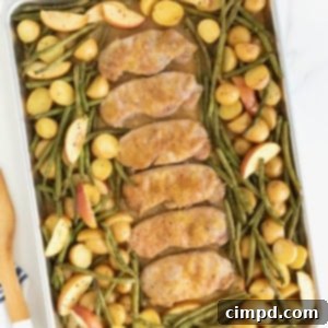 Six pork chops on a rimmed baking sheet surrounded by green beans, apples, and baby potatoes.