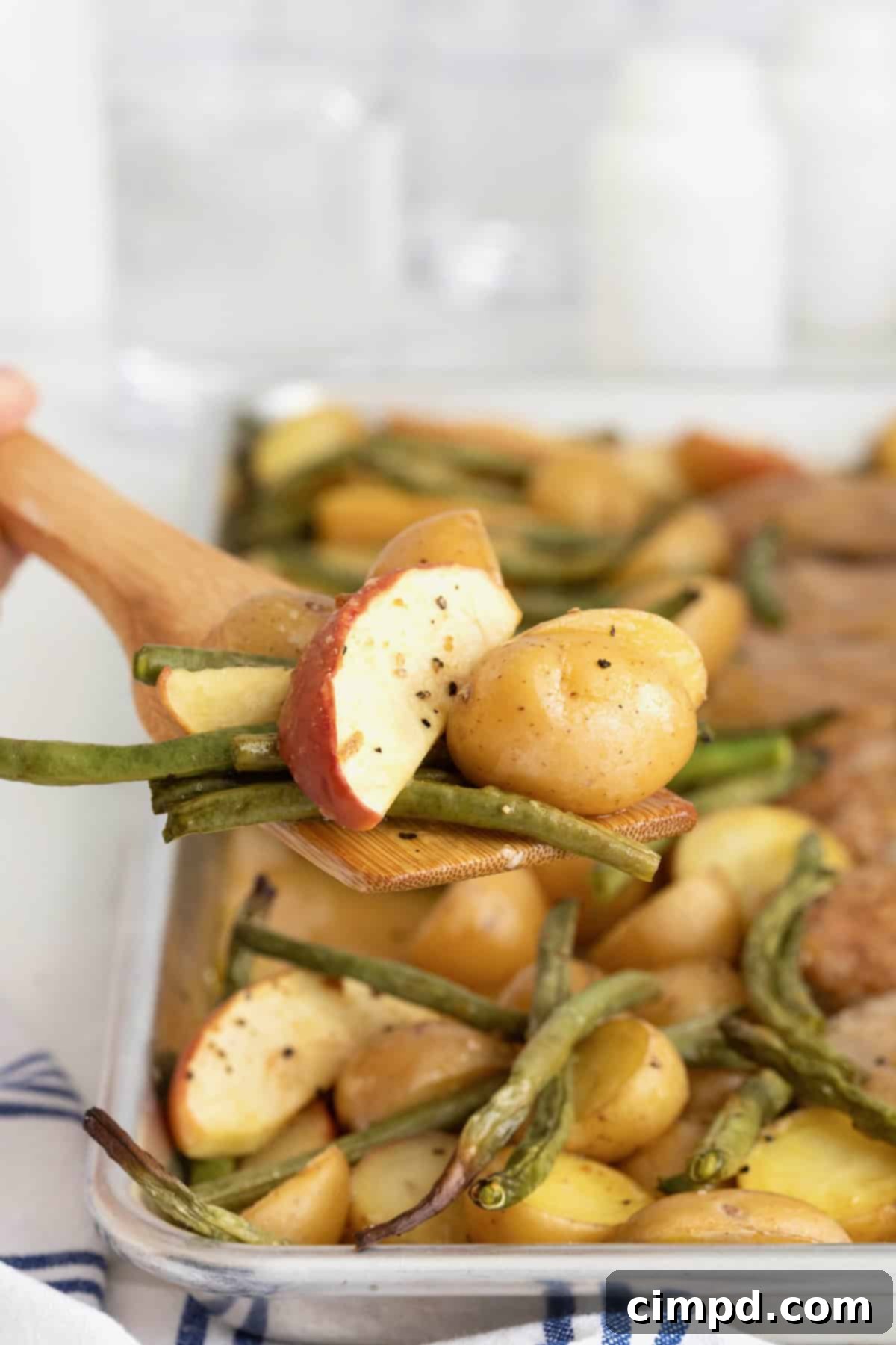 A rustic wooden spatula carefully serving up succulent sheet pan pork chops alongside vibrant green beans, tender apples, and perfectly roasted potatoes from the baking sheet.