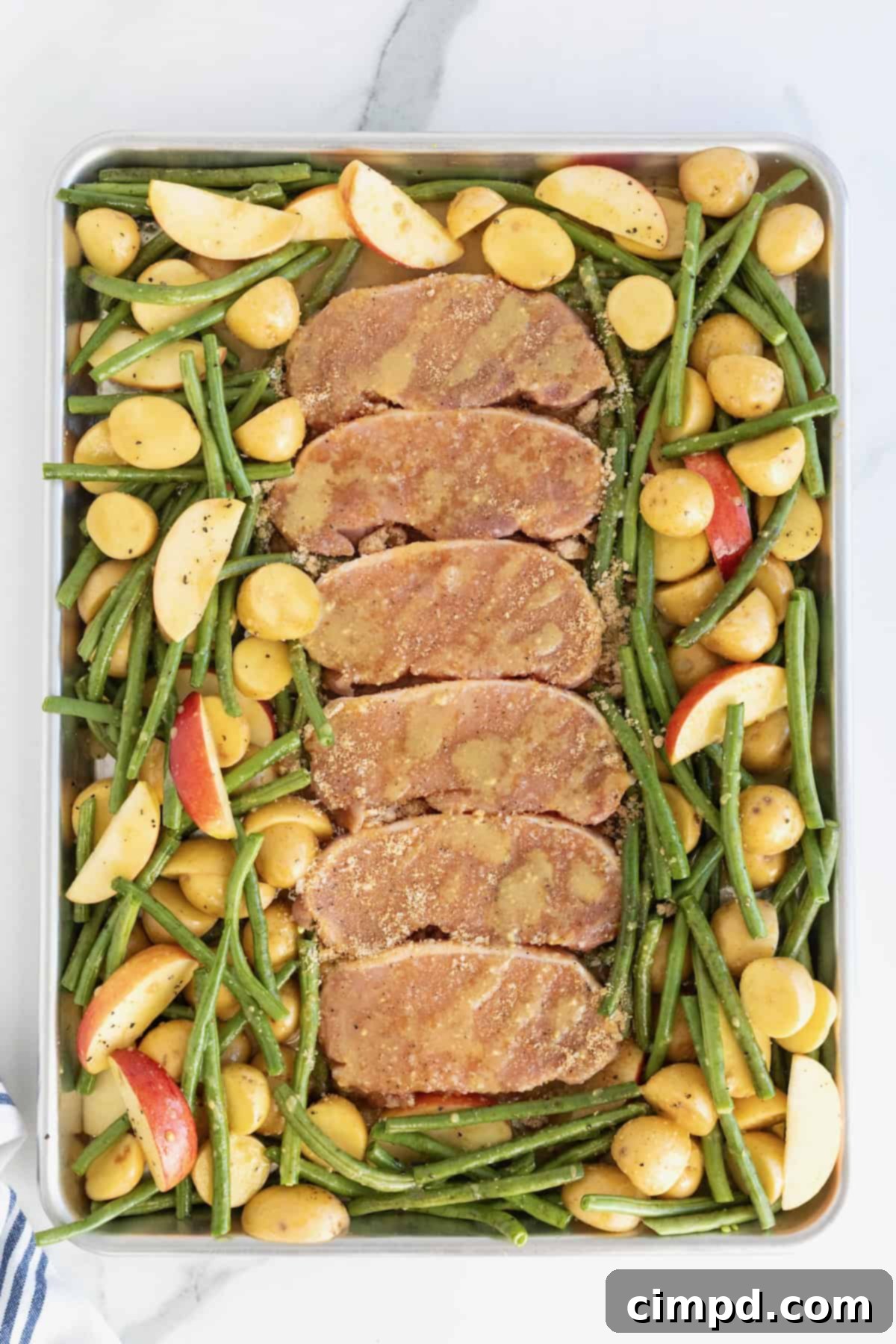 Six seasoned pork chops ready for baking, arranged down the middle of a rimmed baking sheet, generously surrounded by green beans, sliced apples, and halved baby potatoes, all coated in glaze.