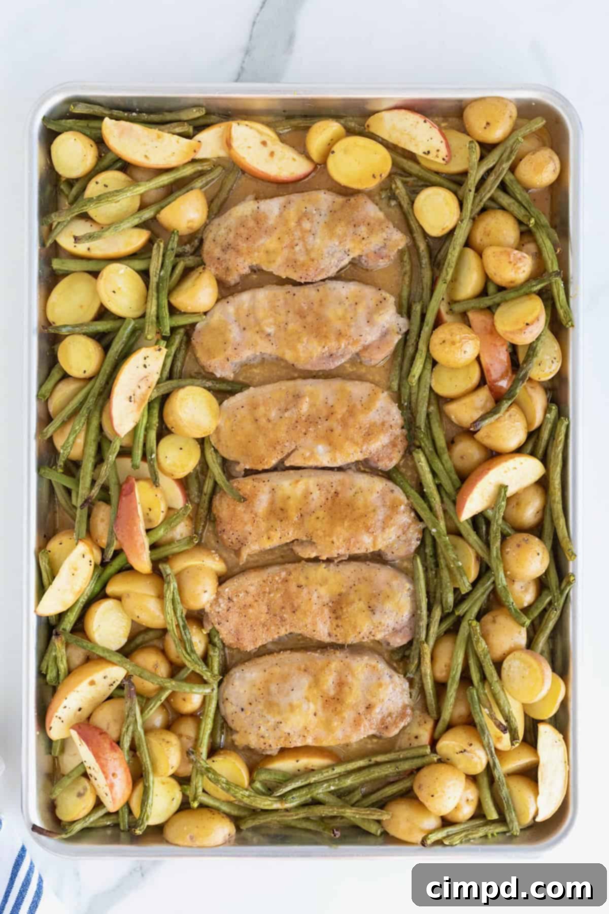Six beautifully baked pork chops, glistening with apple cider glaze, arranged on a rimmed baking sheet. They are perfectly encircled by tender green beans, caramelized apples, and golden roasted baby potatoes.