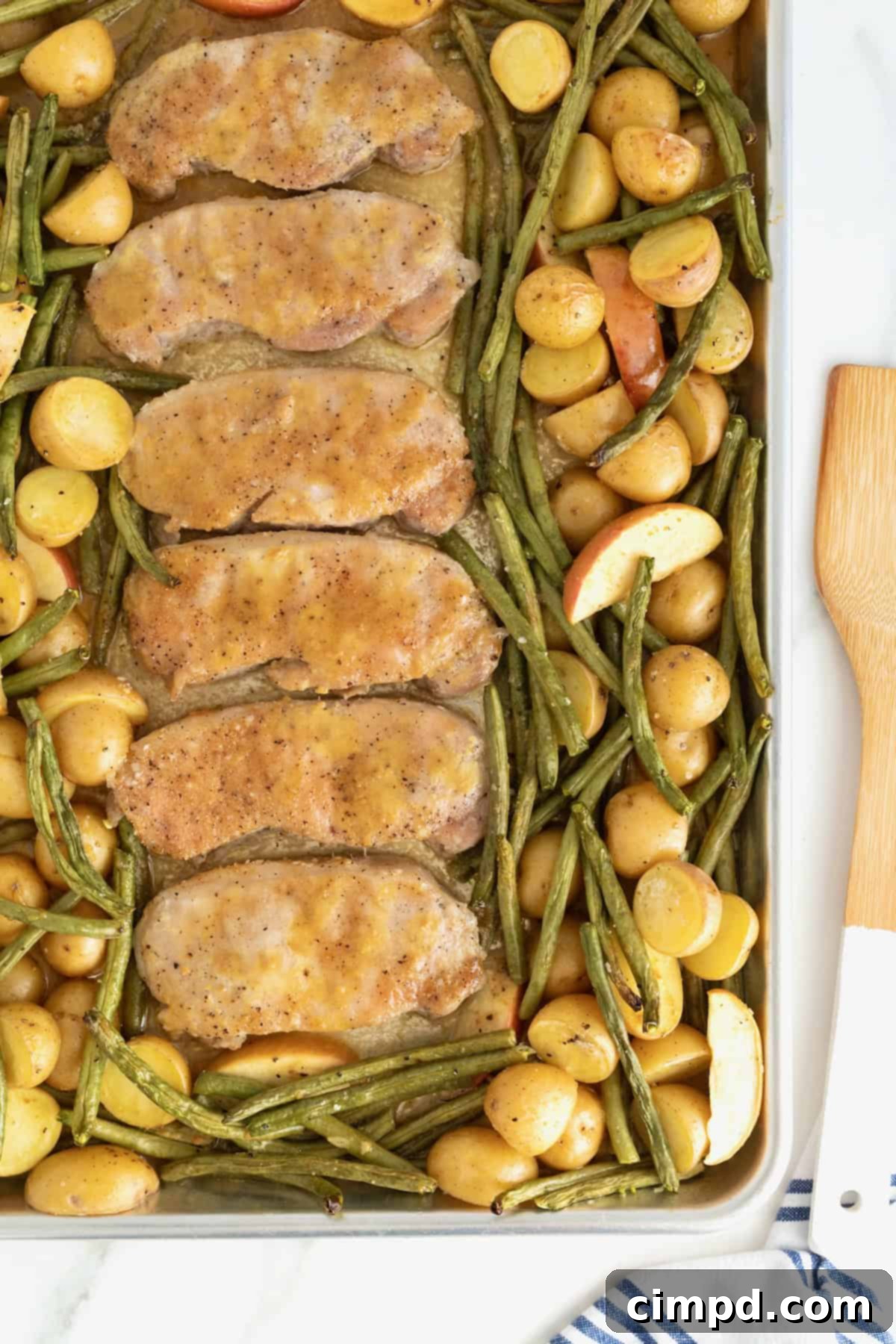 Six tender pork chops, perfectly cooked and coated in glaze, resting on a rimmed baking sheet. They are nestled among vibrant green beans, soft apples, and golden baby potatoes, ready to be served.