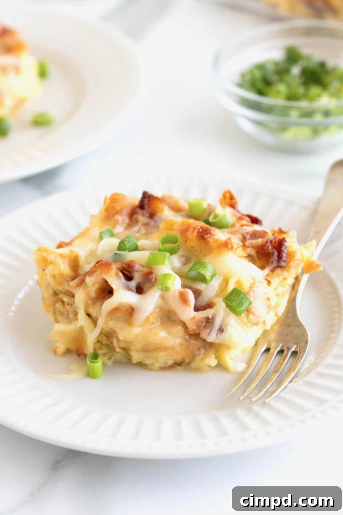 Golden Croissant Breakfast Bake 3 A perfect slice of croissant breakfast casserole served on a white fluted plate with a metal fork, ready to be enjoyed.