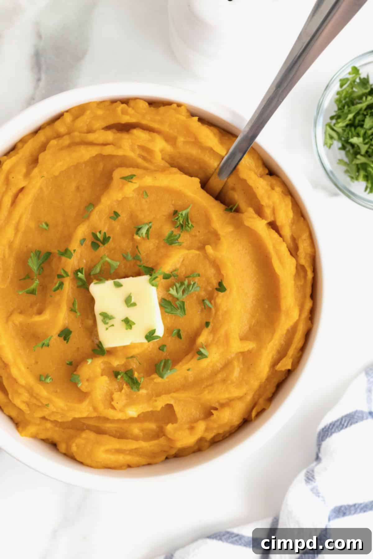 A large white serving bowl overflowing with golden mashed butternut squash, elegantly garnished with a melting square of butter and freshly chopped parsley, ready to be served.