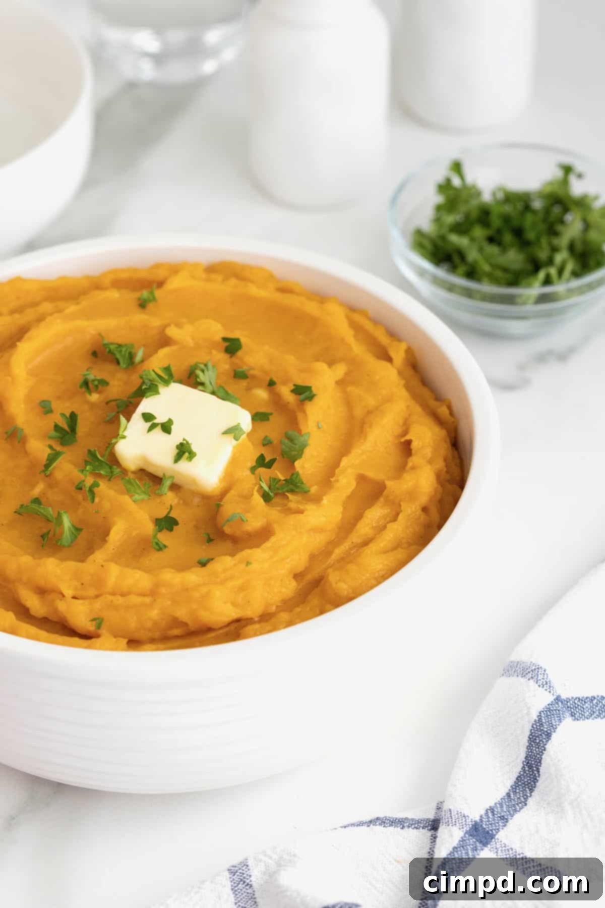 A large white serving bowl showcases golden mashed butternut squash, artfully topped with a square of rich butter and a sprinkle of fresh green parsley, inviting guests to enjoy.