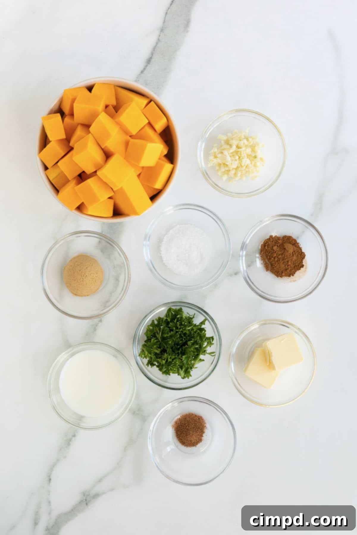 A vibrant display of fresh ingredients for mashed butternut squash, including cubed squash, butter, garlic, brown sugar, and spices, arranged in small glass dishes on a white marble counter.