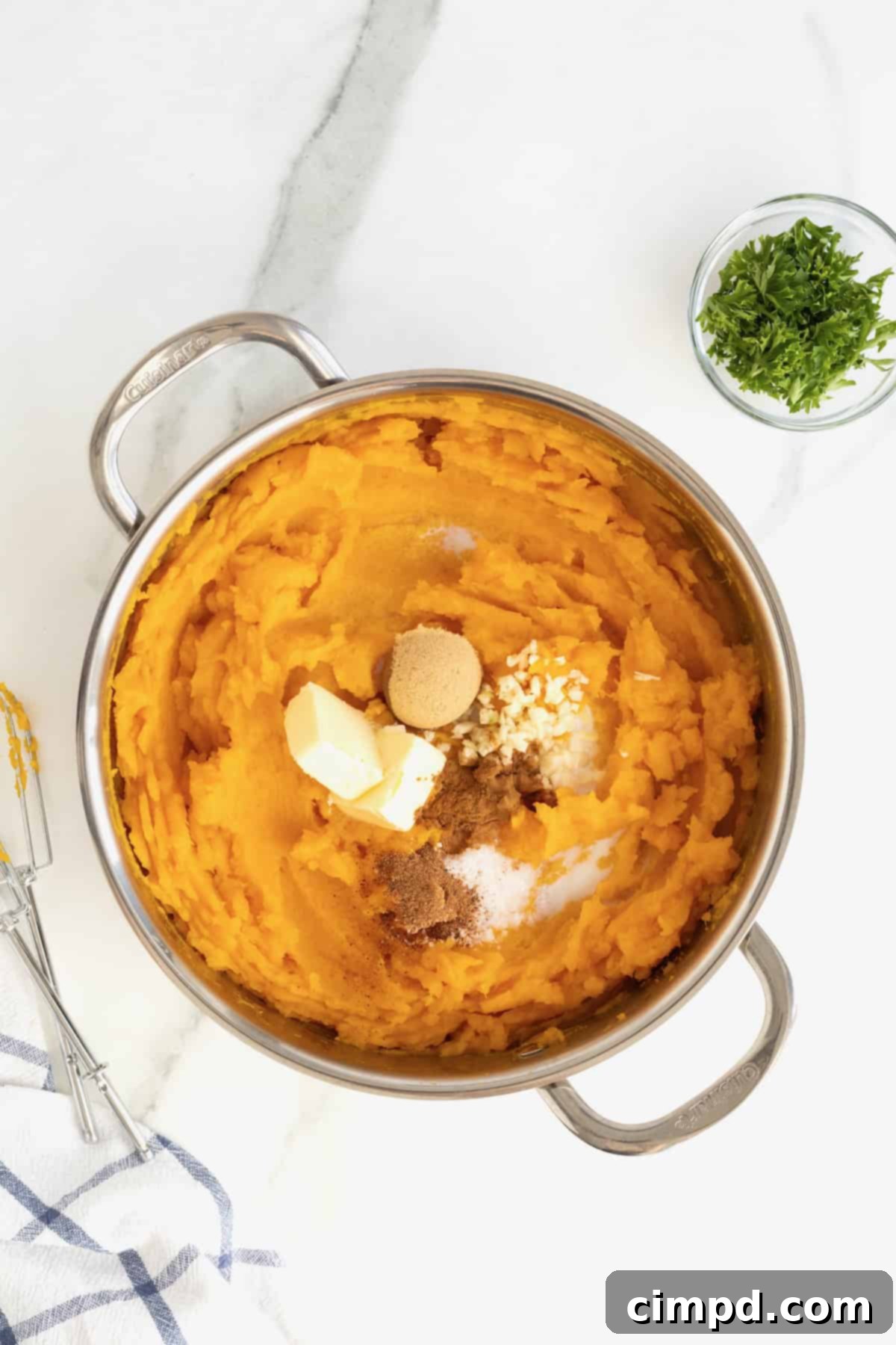 A stainless steel pot containing mashed butternut squash, with fresh butter, brown sugar, minced garlic, cinnamon, nutmeg, and heavy cream added, ready for the final mix.