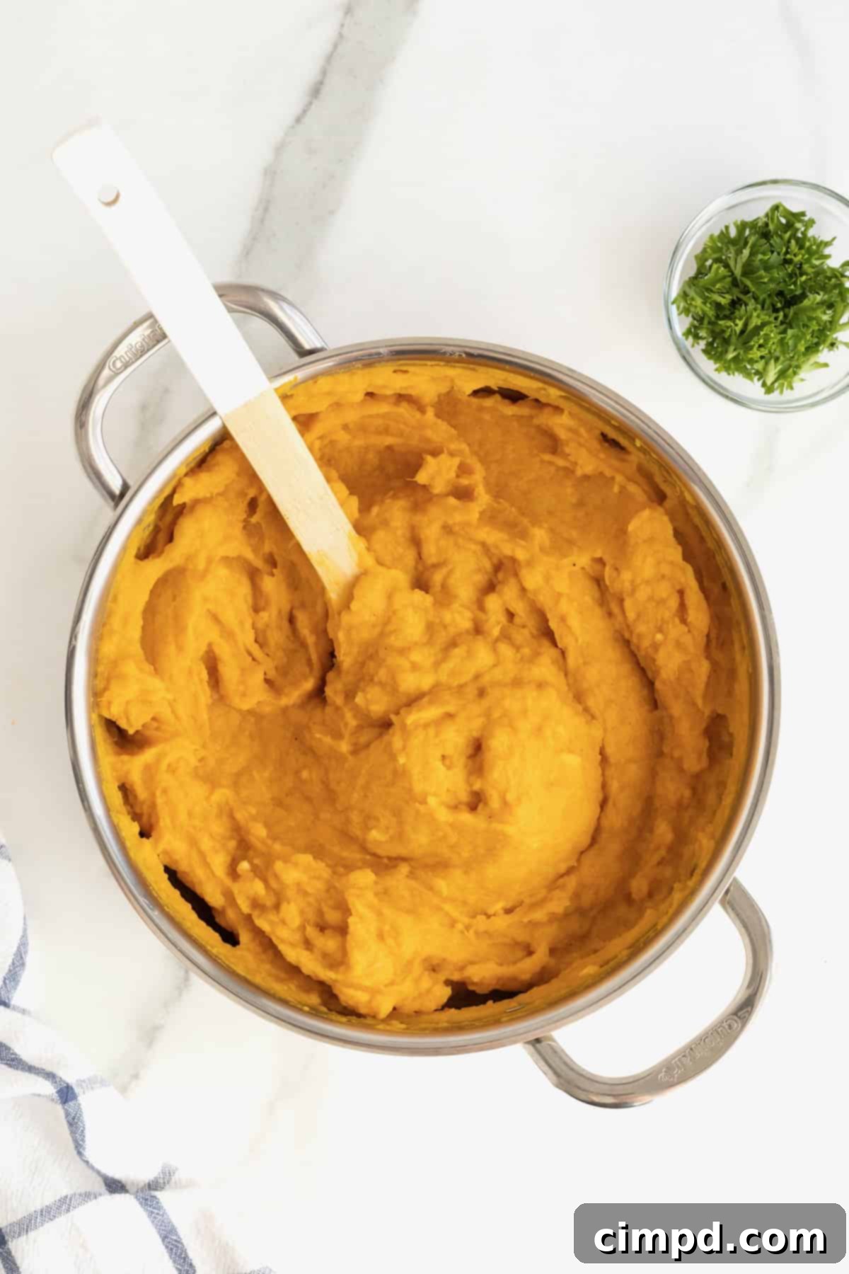 A stainless steel saucepan filled with smooth, creamy mashed butternut squash, featuring a wooden spoon resting in the pot on a white marble counter, indicating its perfect texture.