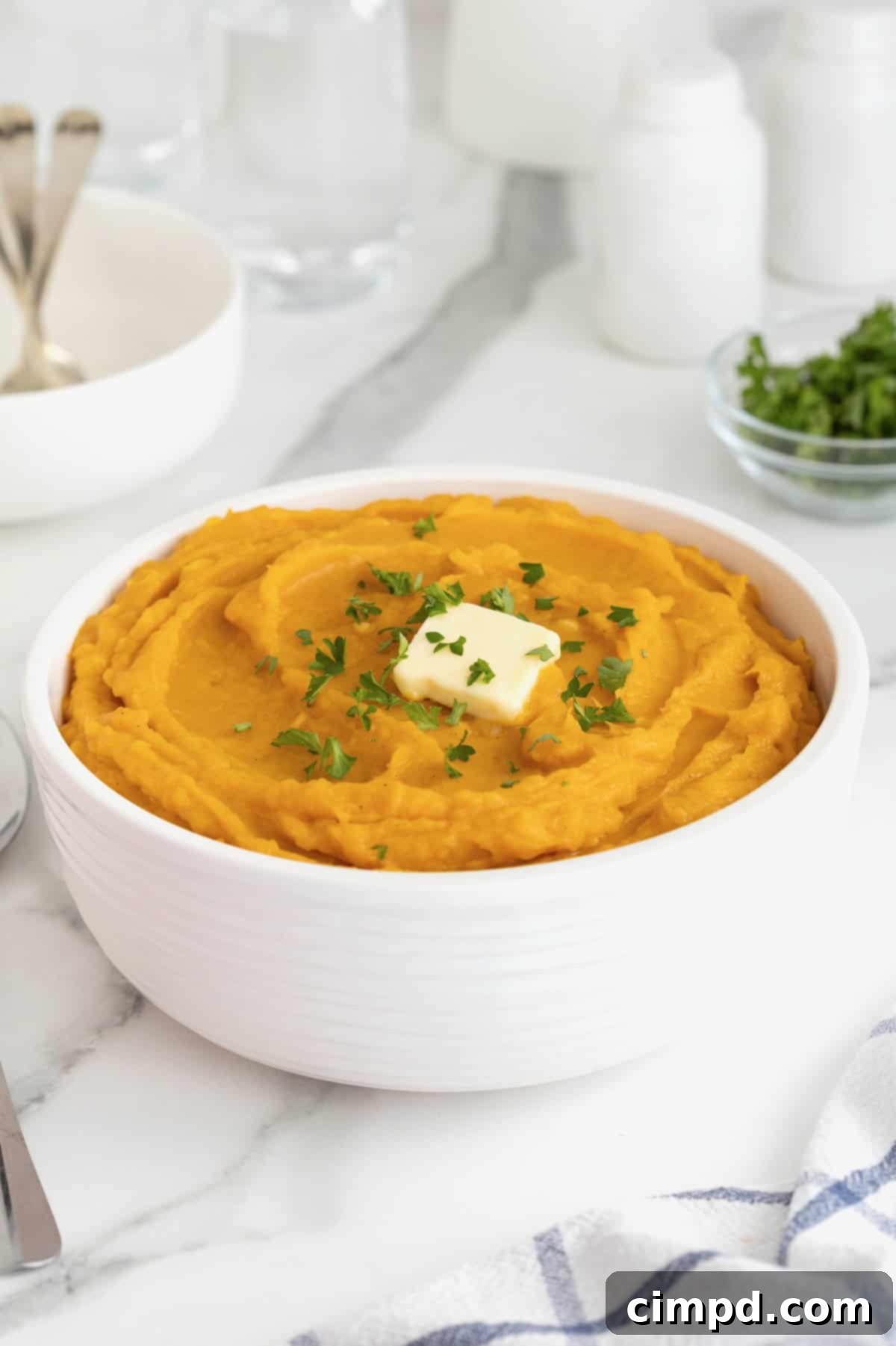 A pristine white serving bowl filled with golden mashed butternut squash, beautifully crowned with a melting cube of butter and a vibrant garnish of fresh parsley, prepared for serving.