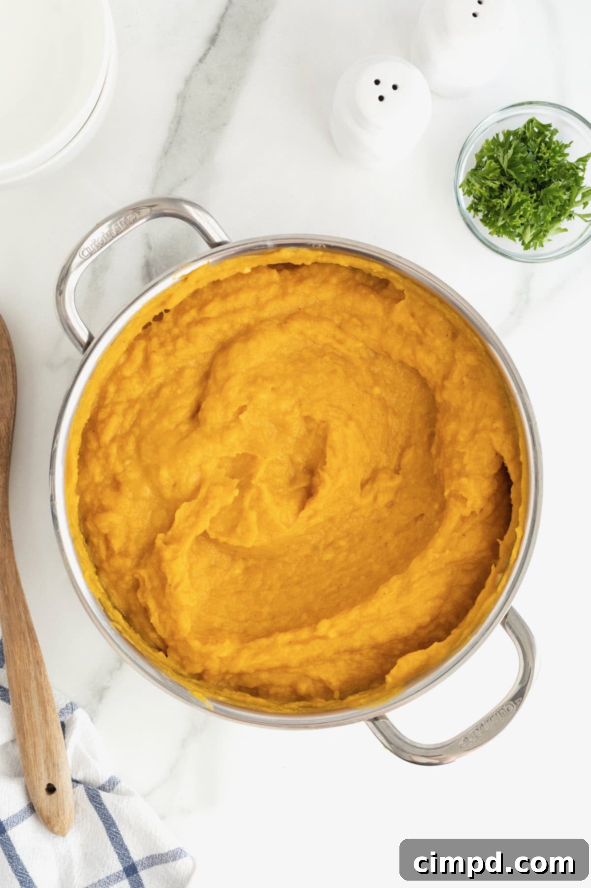 A stainless steel saucepan filled with creamy mashed butternut squash, accompanied by a wooden spoon, set on a bright white marble counter, illustrating a perfectly prepared dish.