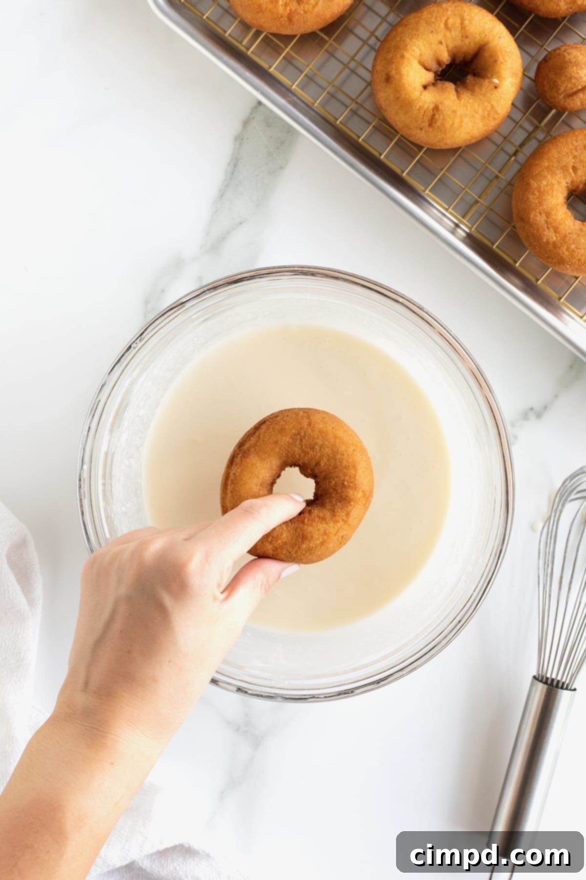 A hand dipping a donut into a glass bowl of icing.
