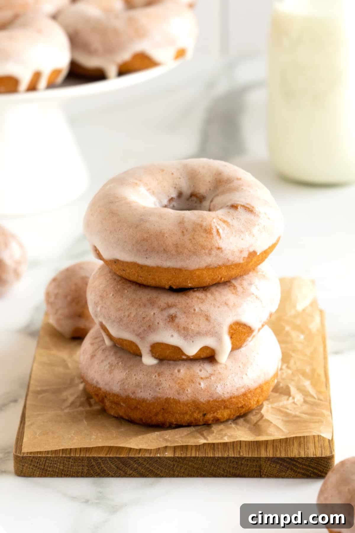 Three glazed homemade donuts stacked on a wooden cutting board.