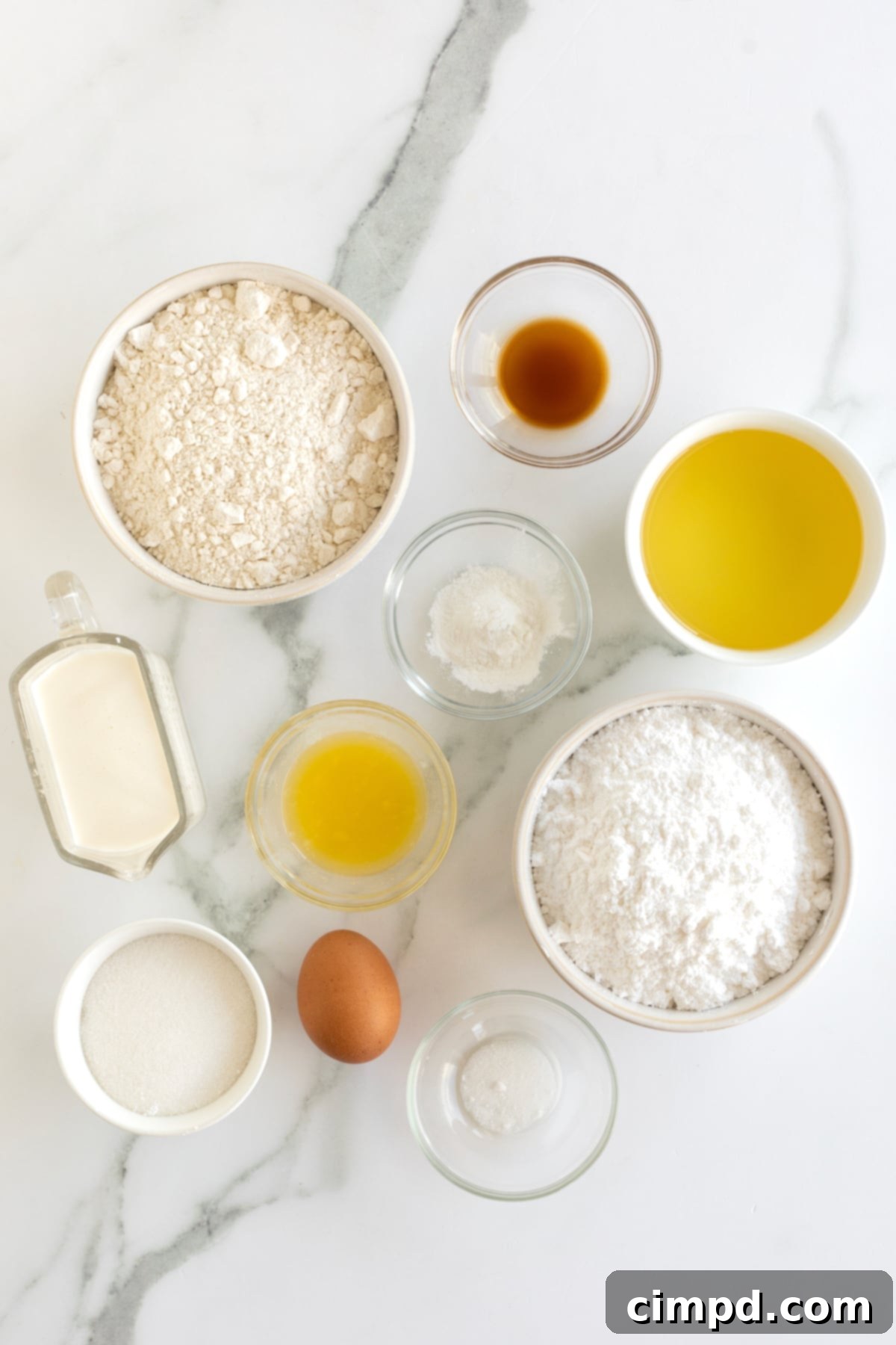 Flour, sugar, melted butter, vanilla extract, heavy cream and canola oil in small glass dishes on a counter next to one brown egg.