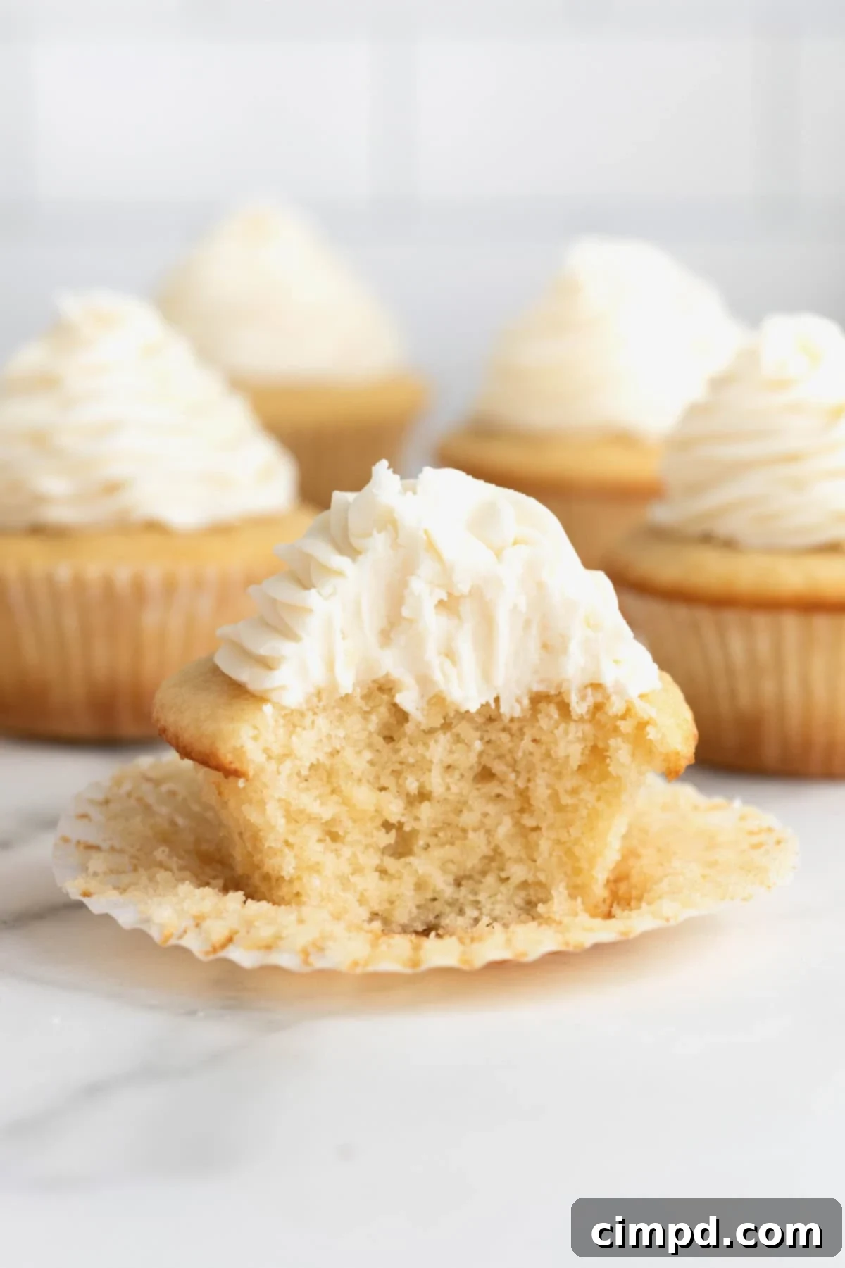 The Perfect Classic Vanilla Cupcake 3 A freshly frosted vanilla cupcake with its paper liner peeled back, revealing a soft, fluffy interior with a bite taken out.