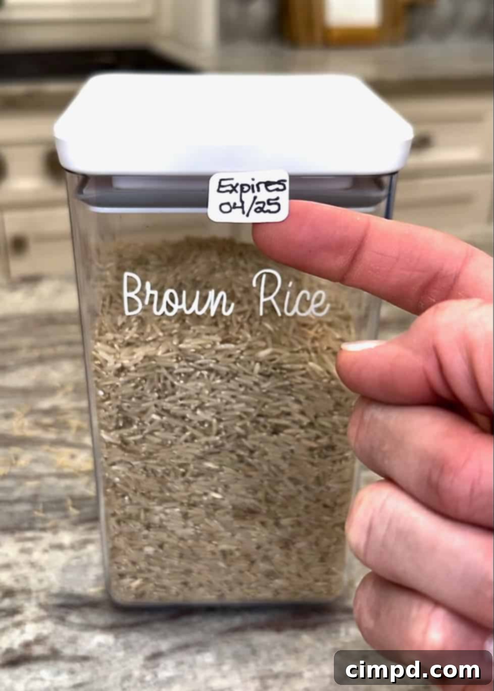 A hand carefully placing a removable label with an expiration date onto a clear canister filled with brown rice in a well-lit pantry.
