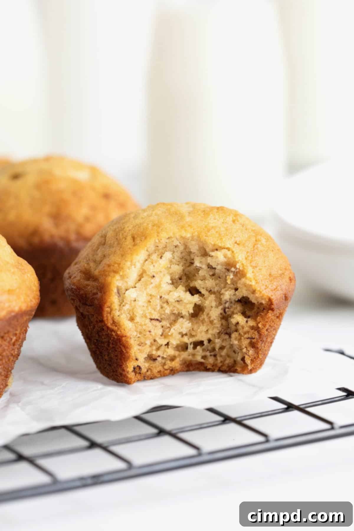 A perfectly baked banana muffin, with a gentle bite taken, resting on a piece of parchment paper on a wire cooling rack.