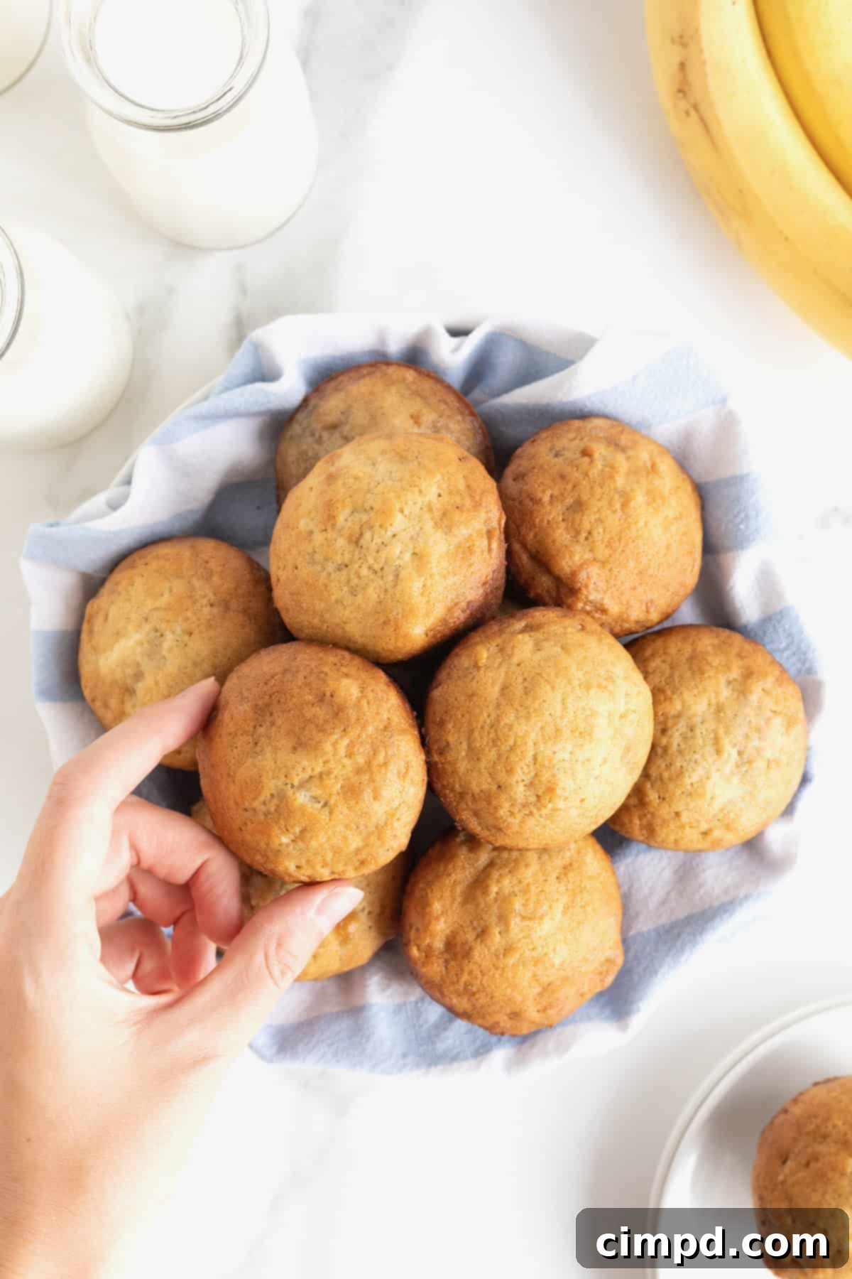 A hand reaching into a large white bowl, lined with a light blue tea towel, to gently pick up one of the freshly baked banana muffins.