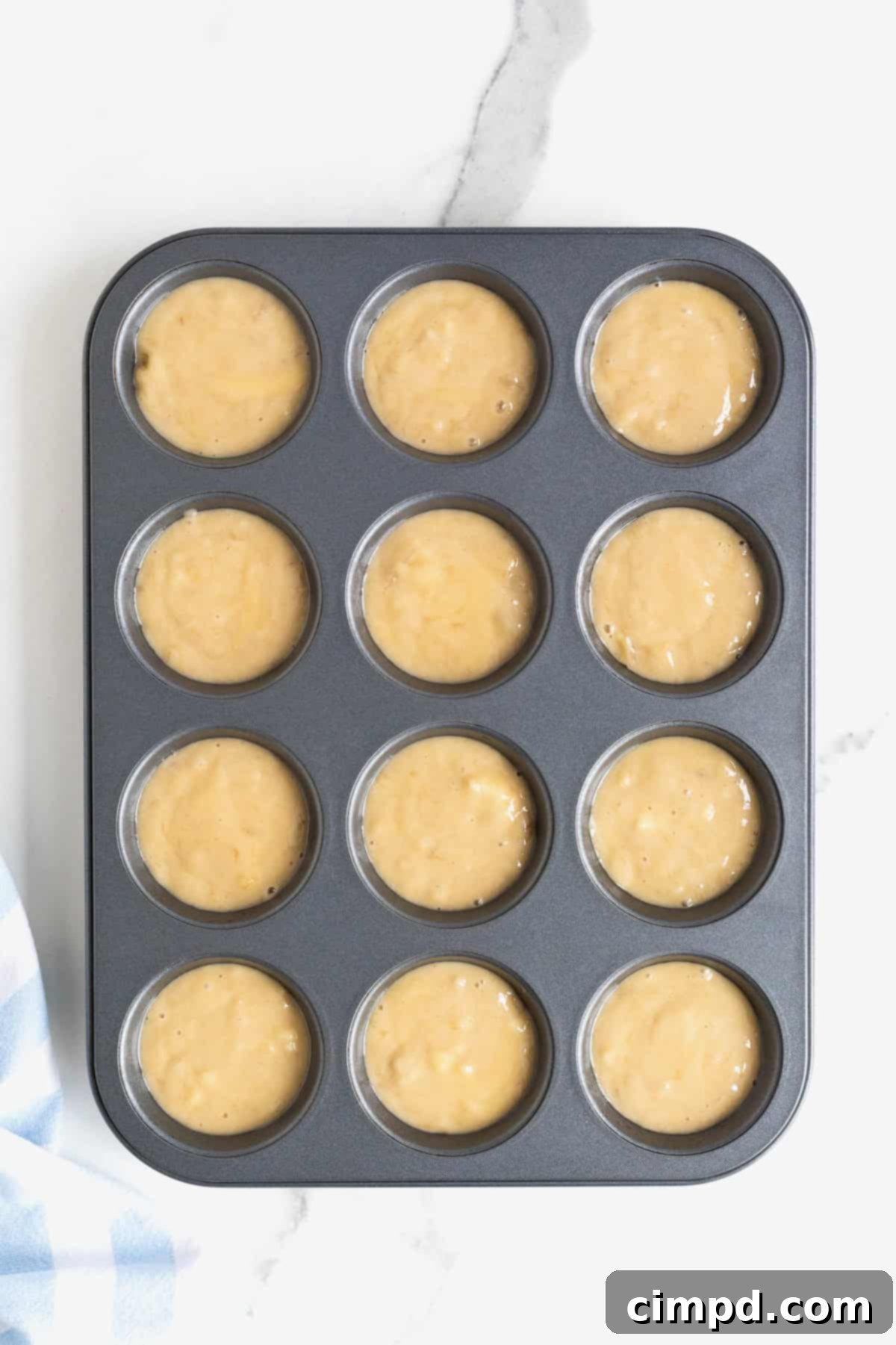 Banana muffin batter neatly scooped into a dark aluminum muffin tin, ready for baking, set on a white marble counter.