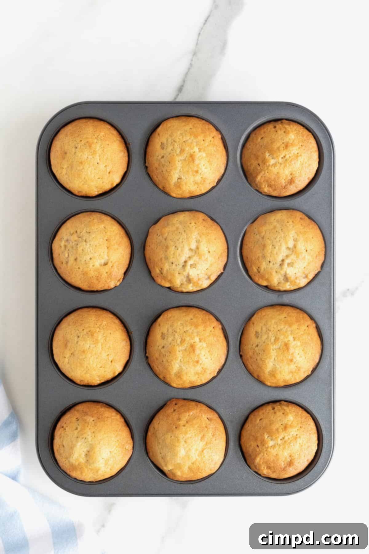 Freshly baked golden banana muffins still in a dark aluminum muffin pan, resting on a white marble counter, emitting a delicious aroma.