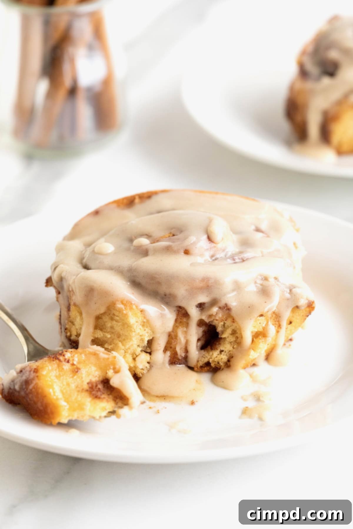 A cinnamon roll covered in icing on a small white plate with a fork. 