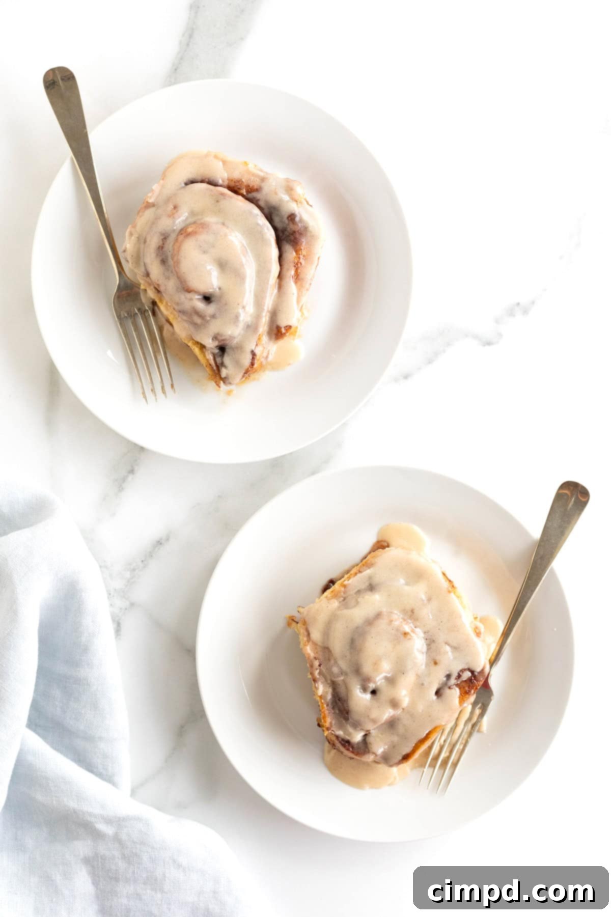 Two cinnamon rolls covered in icing on  small white plates with forks.