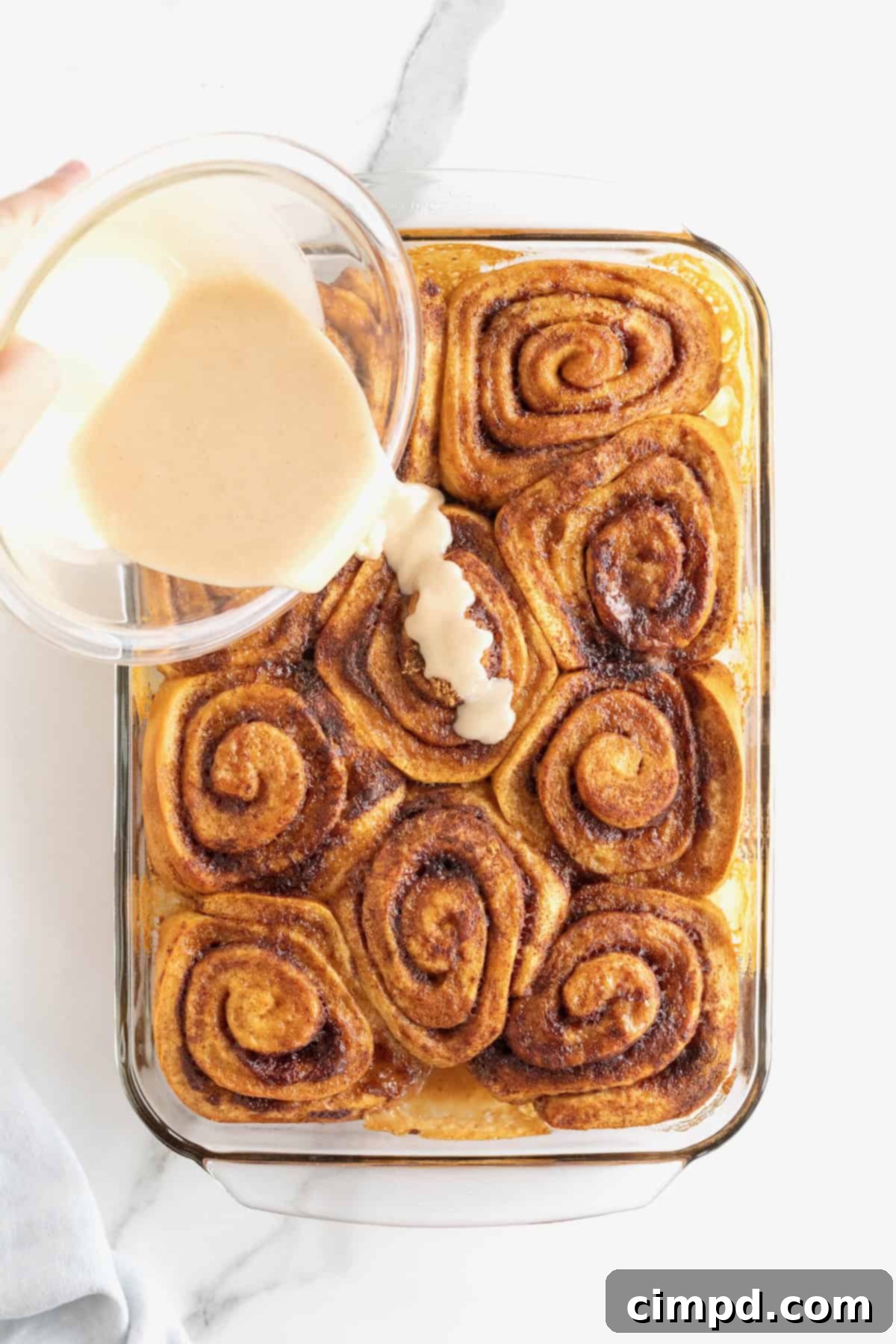Cinnamon vanilla icing being poured over baked cinnamon rolls in a glass baking dish.