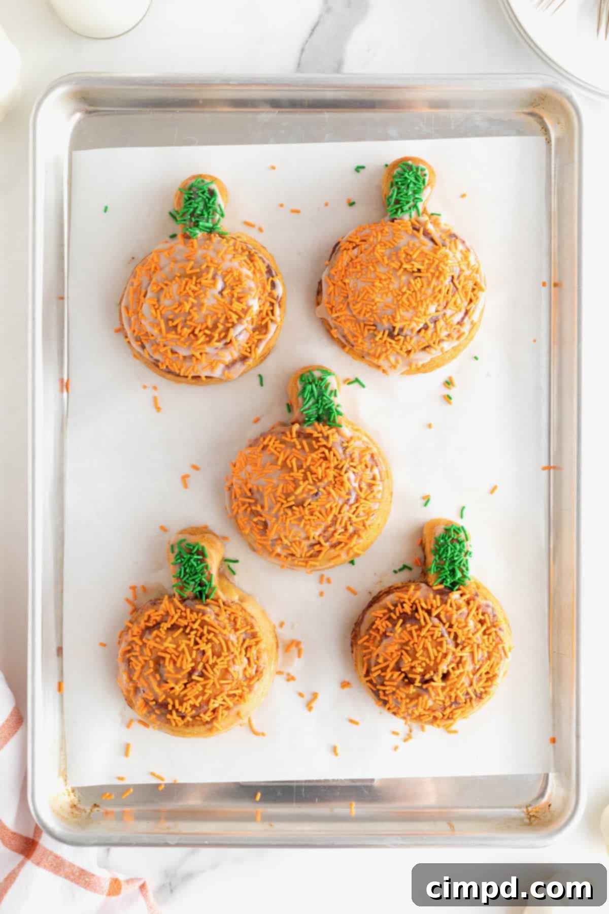Five frosted pumpkin shaped cinnamon rolls with orange and green sprinkles on a parchment lined aluminum baking sheet.