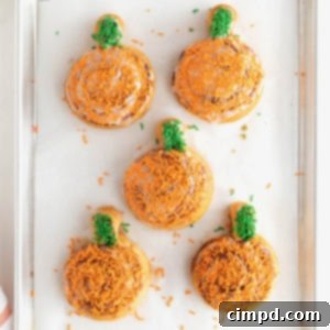 Five frosted pumpkin shaped cinnamon rolls with orange and green sprinkles on a parchment lined aluminum baking sheet.
