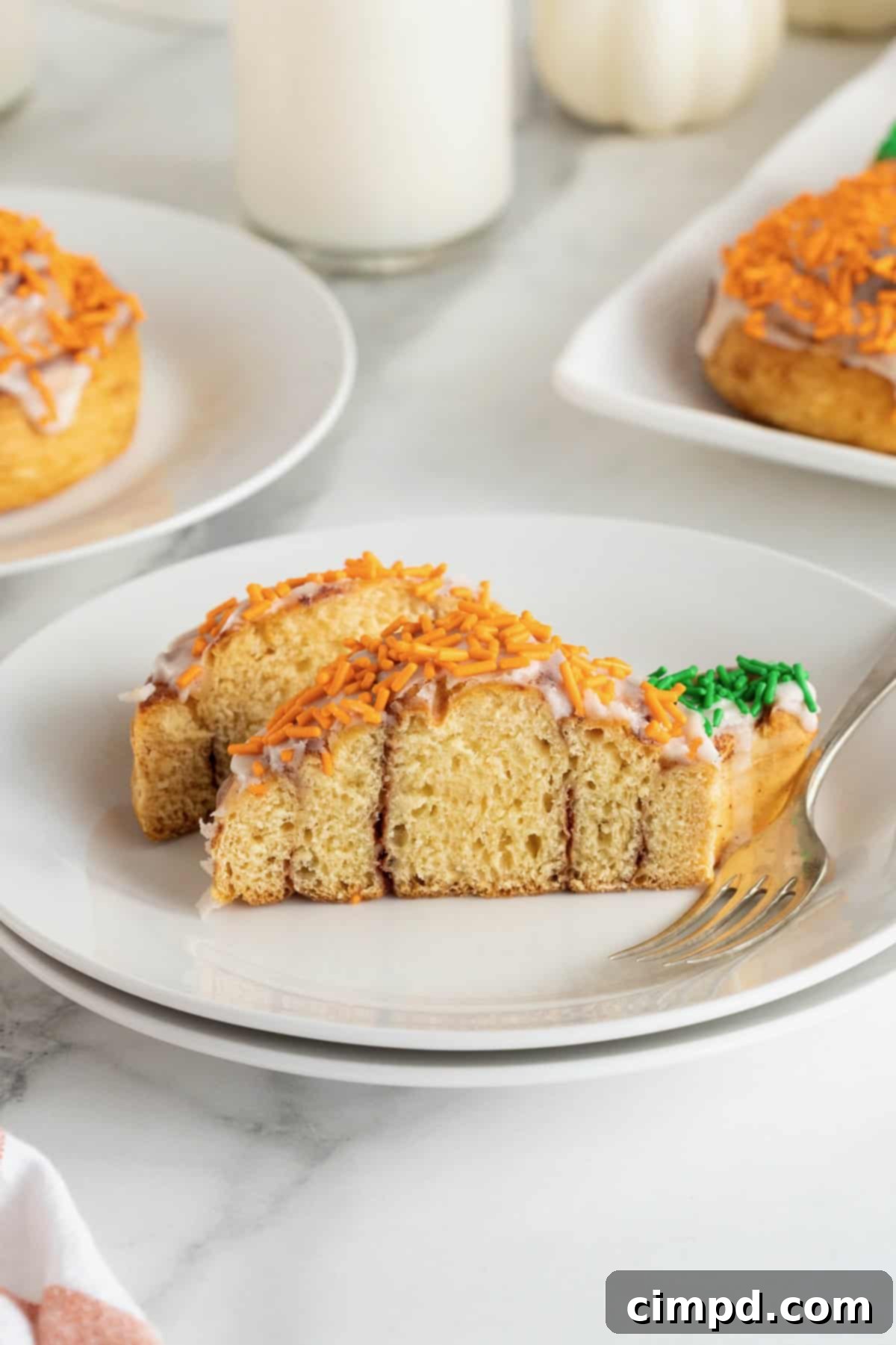 A pumpkin shaped cinnamon roll cut in half with a fork resting beside it on a small white plate.