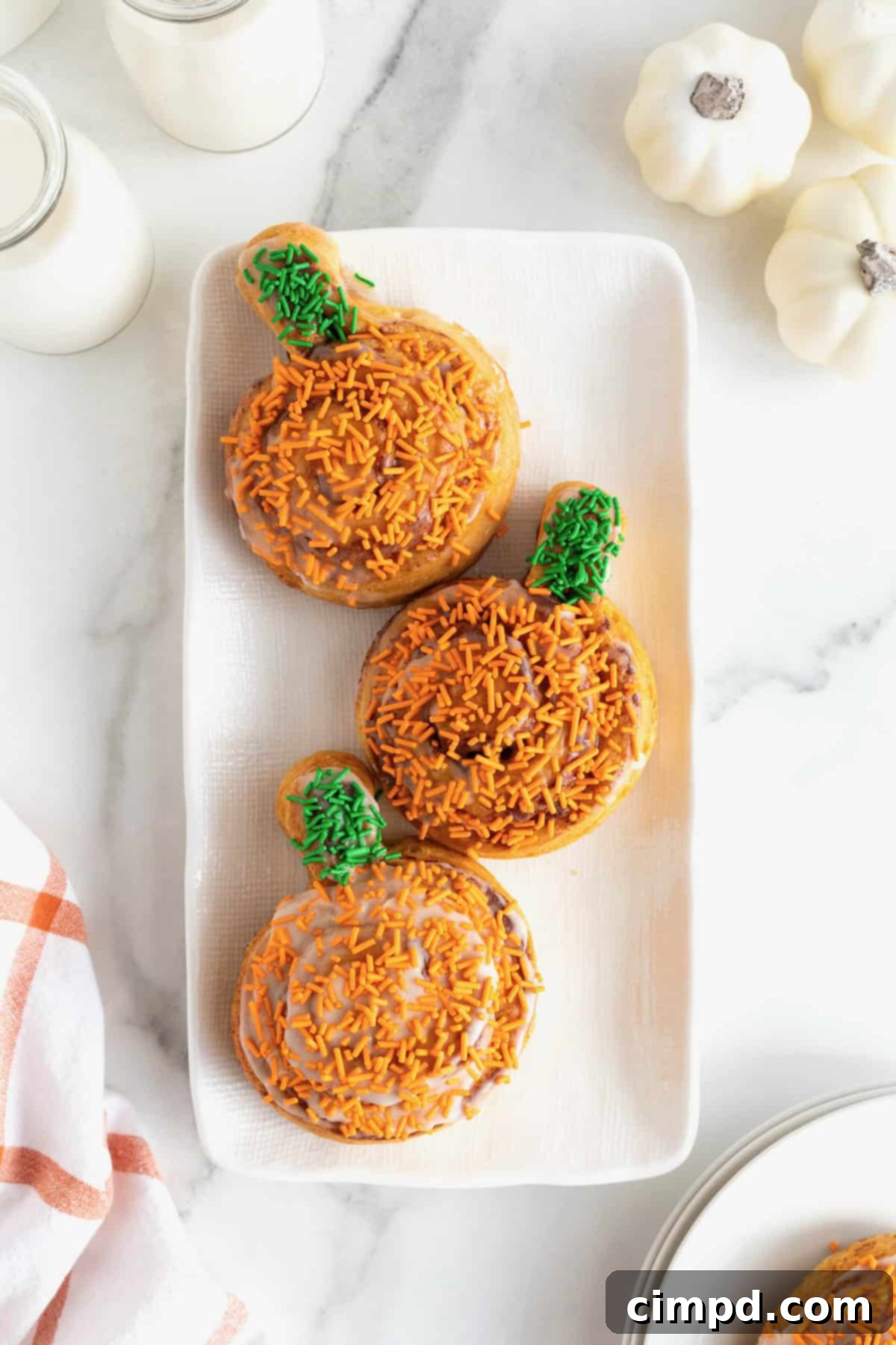 Three cinnamon rolls shaped like pumpkins with orange and green sprinkles on a rectangular white serving tray.