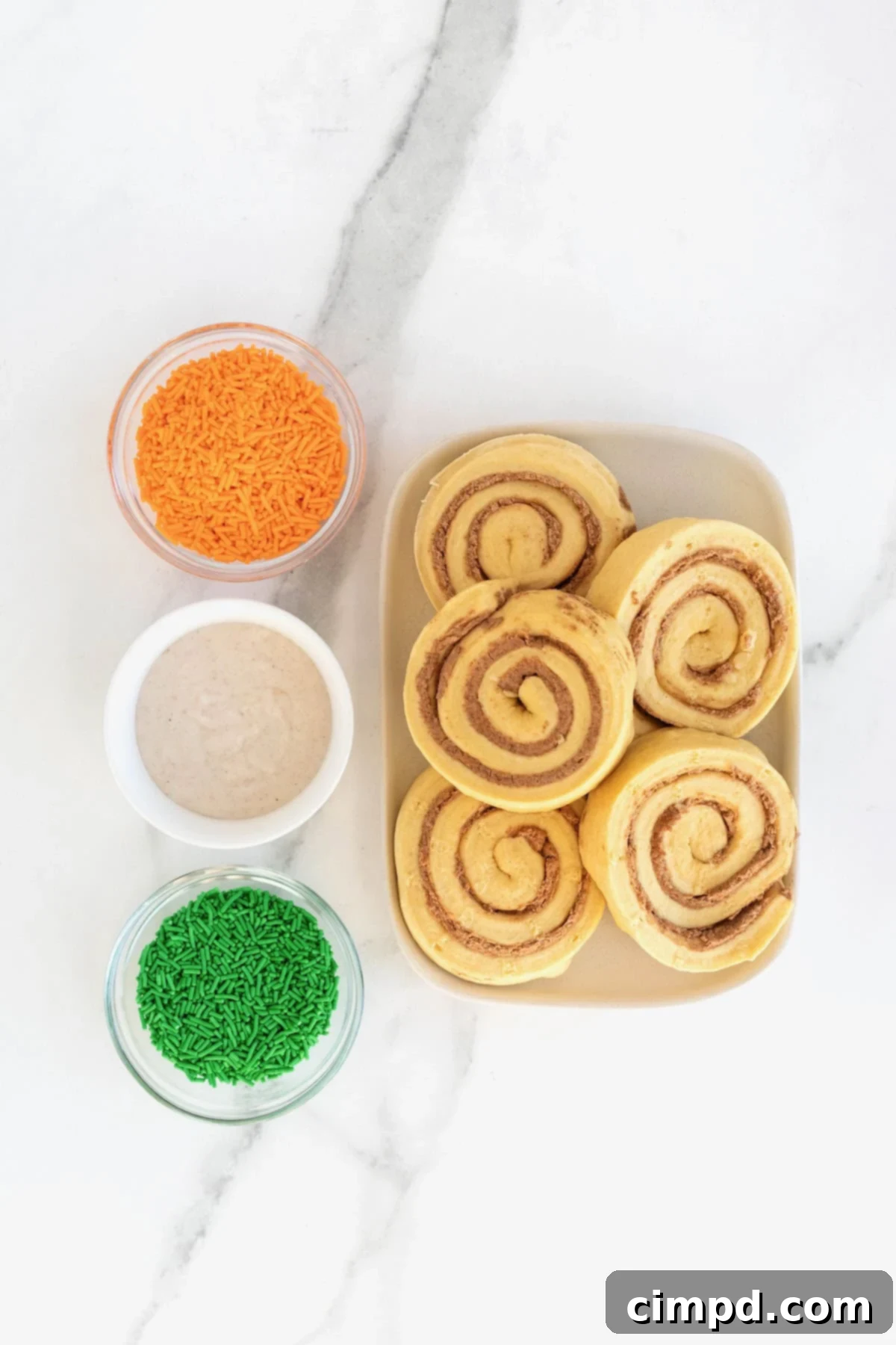Ingredients to make pumpkin shaped cinnamon rolls in white dishes on a white marble counter.