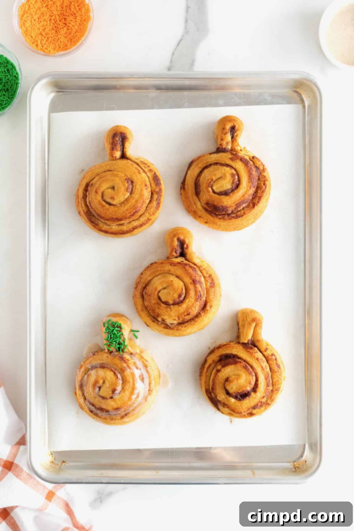 Five iced pumpkin cinnamon rolls on a parchment lined aluminum baking sheet. One of the pumpkins has  green sprinkles on the stem.