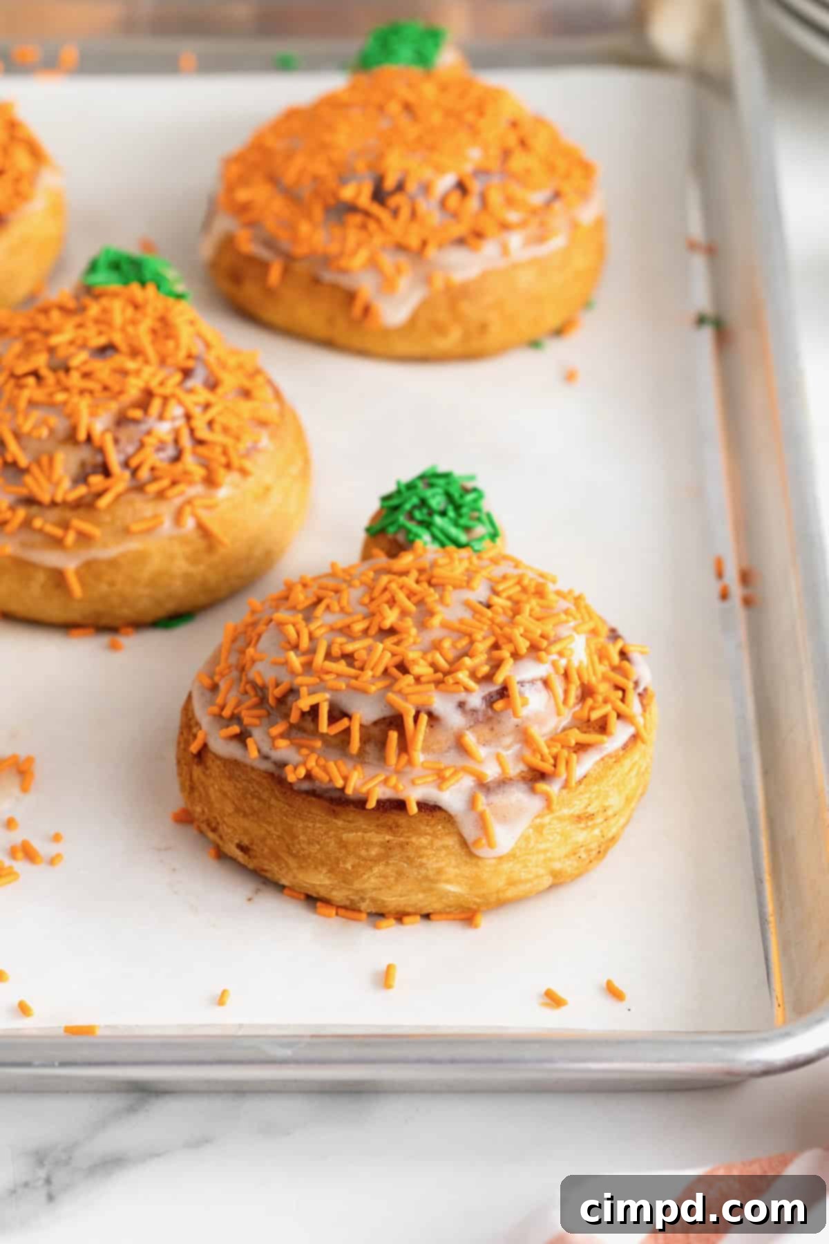 Iced pumpkin shaped cinnamon rolls with orange and green sprinkles on a parchment lined aluminum baking sheet.
