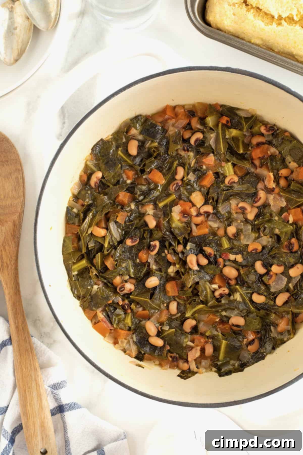 A cream colored Dutch oven filled with simmering black eyed peas and collard greens, placed on a pristine white marble counter. A wooden spoon rests invitingly beside the pot, ready to serve.