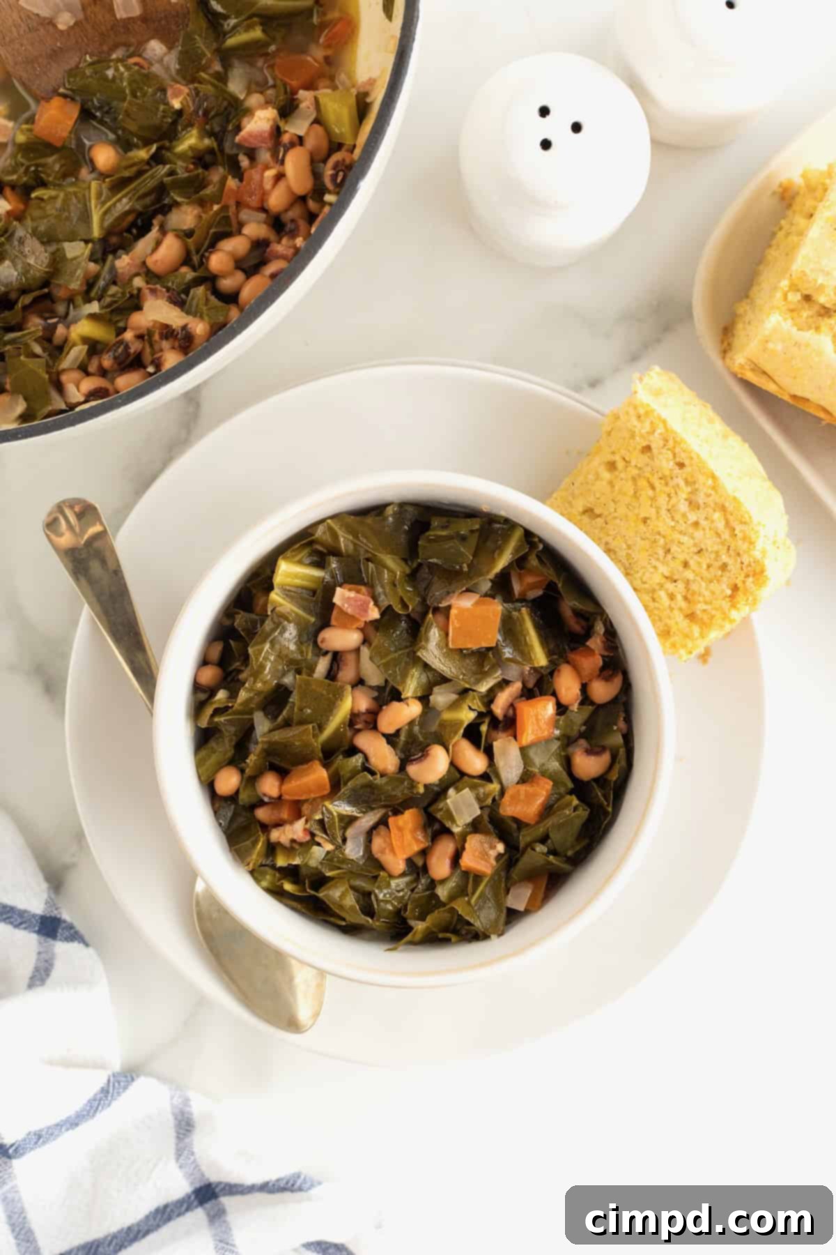 A white soup bowl filled with perfectly cooked black eyed peas and collard greens, presented beautifully on a white plate with a golden, square piece of cornbread and an elegant spoon beside it. 