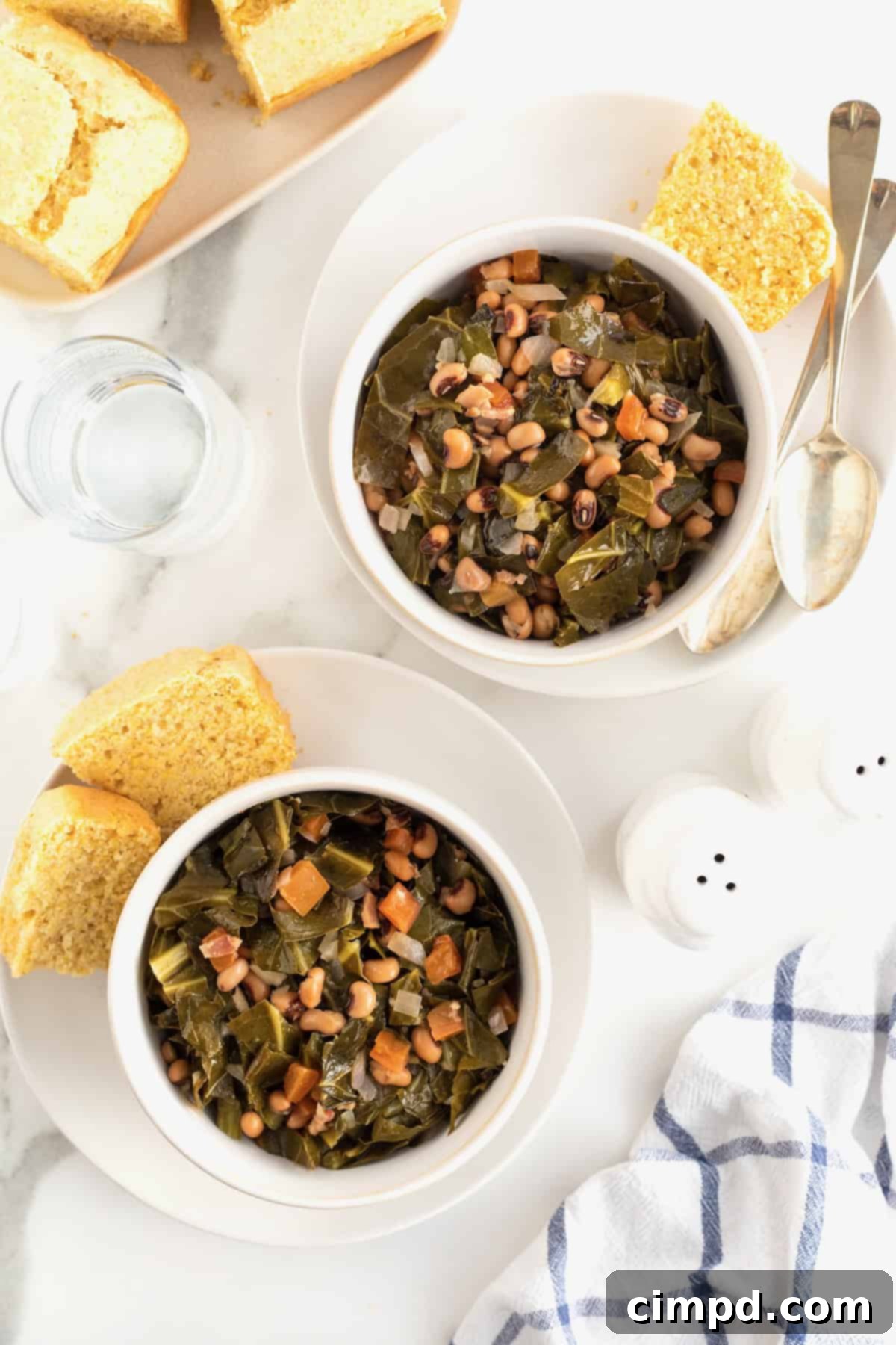 Two pristine white bowls filled with perfectly cooked black eyed peas and tender collard greens, each garnished and served with a golden chunk of cornbread, resting elegantly on white plates.