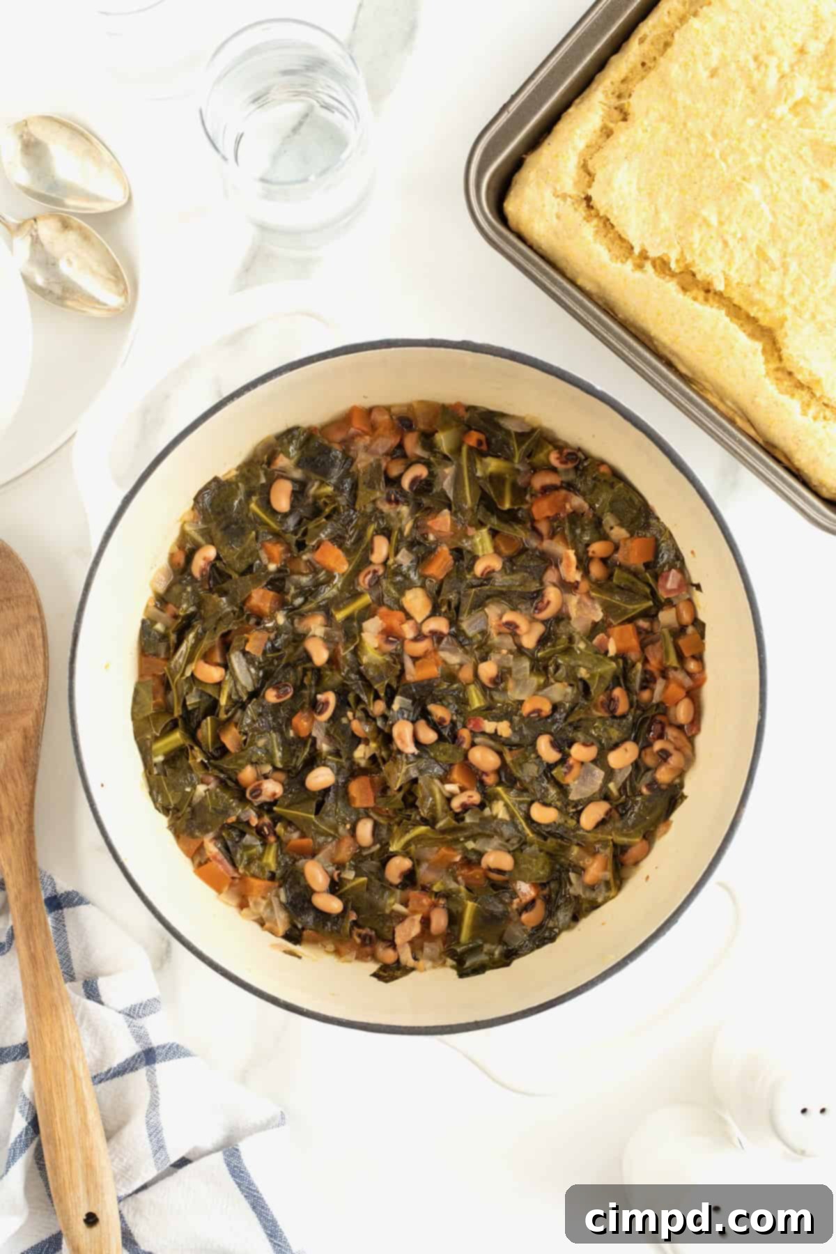 A large, rustic pot of black eyed peas and collard greens resting invitingly next to a golden, freshly baked pan of cornbread on a bright white marble counter, creating a tempting display of heartwarming comfort food.