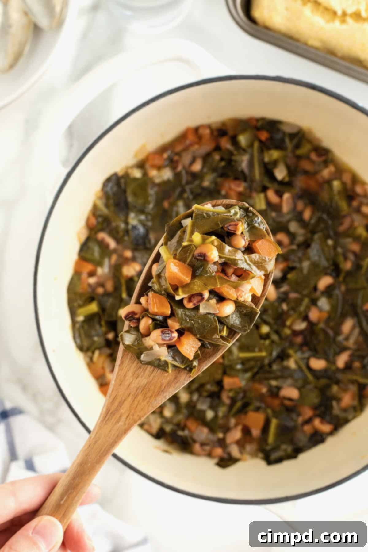 A large white Dutch oven brimming with luscious black eyed peas and collard greens, with a wooden spoon gently lifting a serving, showcasing the rich, comforting texture of the traditional dish.