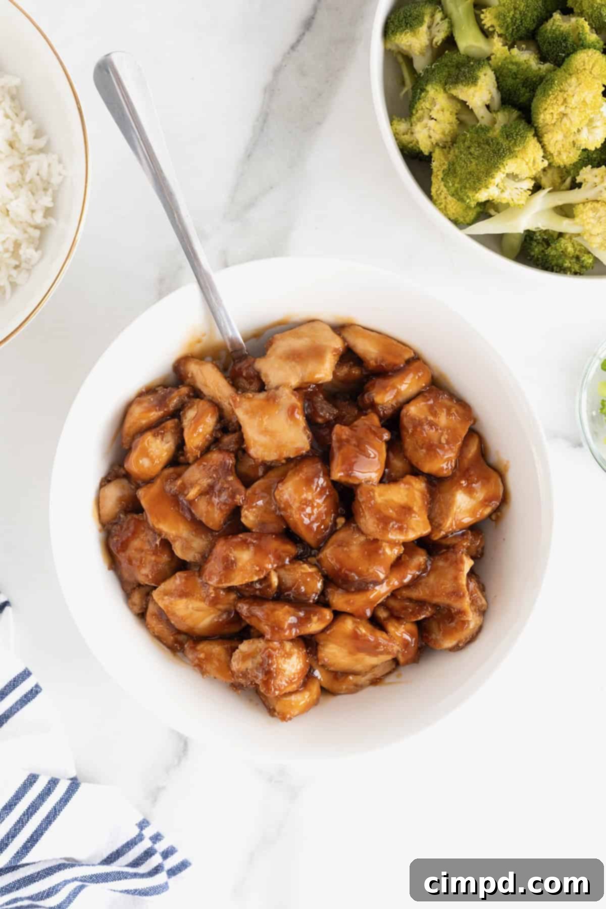 Chunks of chicken in a rich brown sauce, garnished with fresh green onions, served in a pristine white bowl on a elegant white marble countertop, highlighting the dish's vibrant appeal.