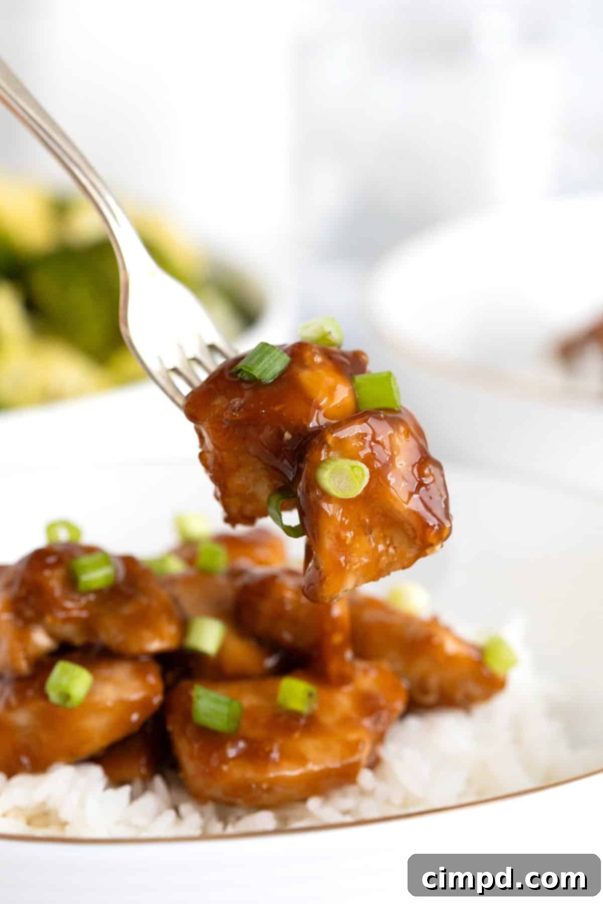 A fork holding two glistening chunks of baked orange chicken, lightly sprinkled with fresh green onion, positioned against a blurred background of a full dish of orange chicken and fluffy white rice.