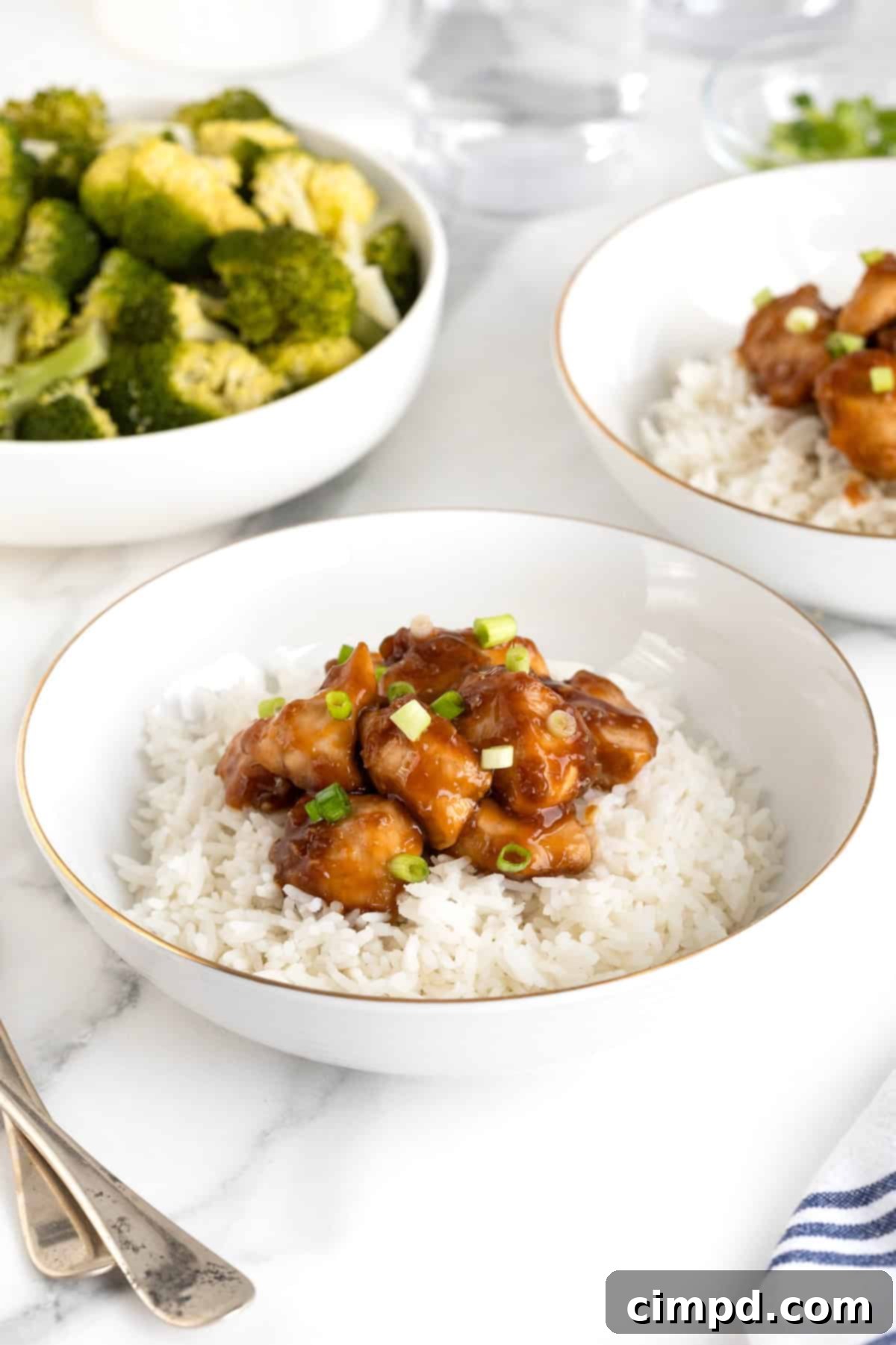 A beautifully presented white and gold-rimmed bowl filled with fluffy white rice, generously topped with baked orange chicken. A side of vibrant green broccoli florets sits artfully in the background, completing a balanced and appetizing meal.