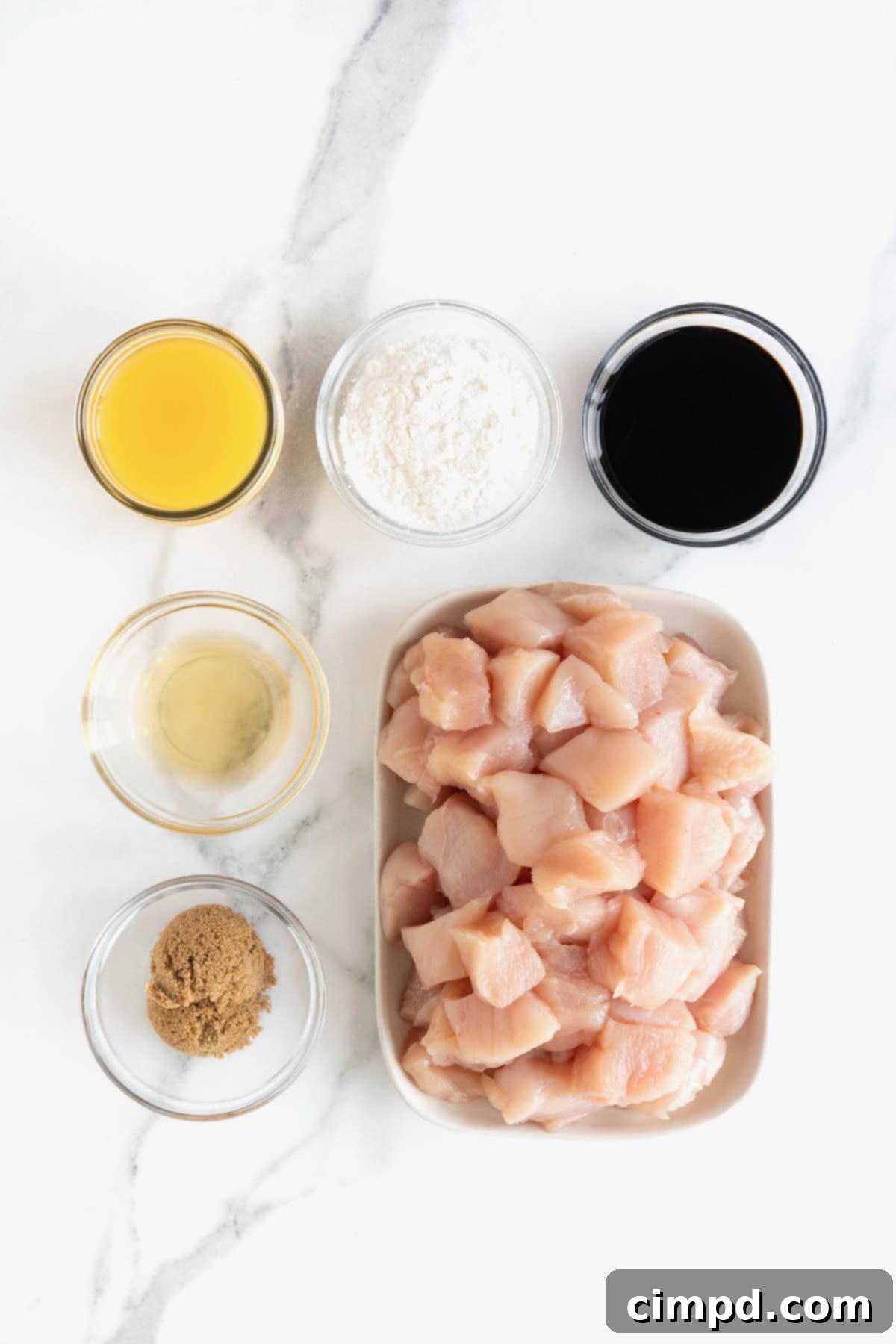 Various ingredients for baked orange chicken, including cubed chicken, cornstarch, soy sauce, orange juice, brown sugar, and rice vinegar, neatly arranged in small glass dishes on a pristine white marble counter, ready for preparation.