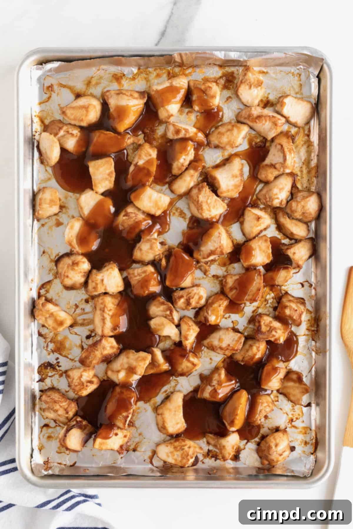Golden-brown chunks of baked chicken, generously coated in a rich, glossy brown orange sauce, spread out on a rimmed baking sheet, ready for the final glaze.