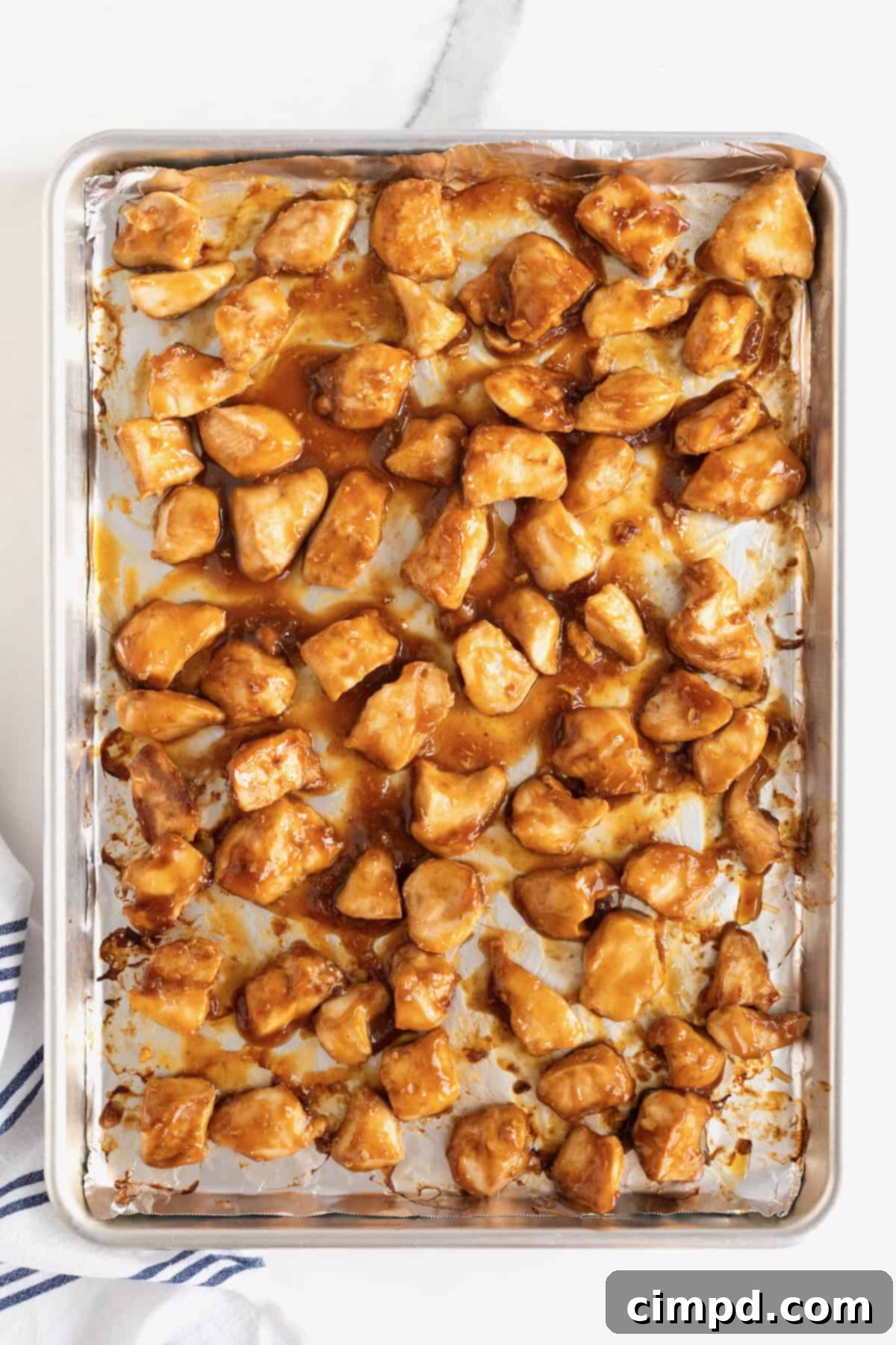 A close-up shot of perfectly baked orange chicken, glistening with a rich, caramelized sauce, spread evenly on a rimmed baking sheet, fresh out of the oven and ready to be served.