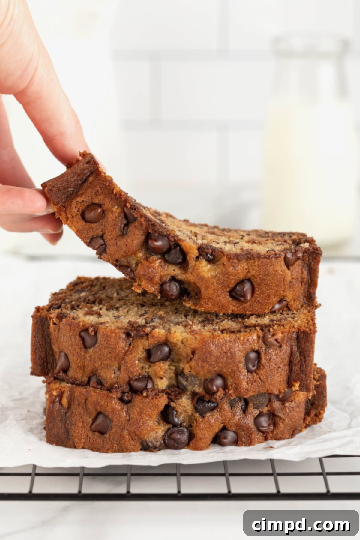 Three golden-brown slices of homemade chocolate chip banana bread, stacked artfully on a cooling rack. A hand reaches in to lift the top slice, revealing the moist interior and abundant melted chocolate chips.